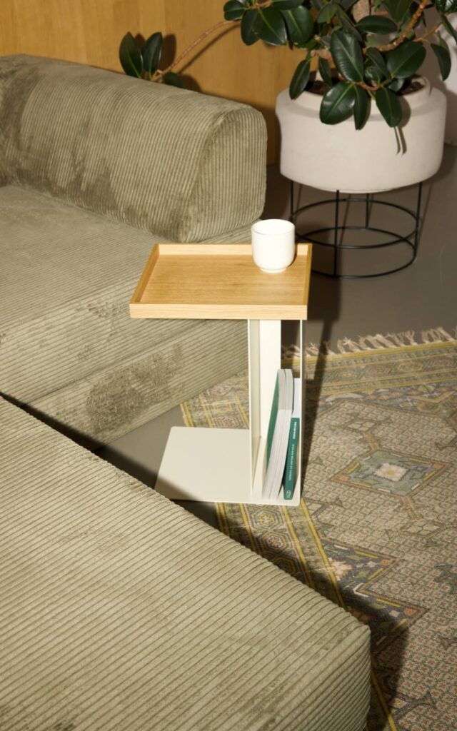 Ein kleiner moderner Beistelltisch mit einer hölzernen Ablagefläche hält eine weiße Tasse. Darunter werden Zeitschriften aufbewahrt. Der Tisch steht zwischen zwei grünen Kordsofas, neben einer Topfpflanze und einem gemusterten Teppich.