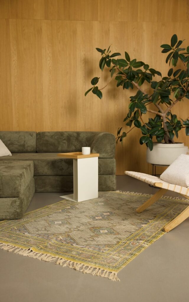 Ein modernes Wohnzimmer mit einem grünen Samtsofa, einem weißen Beistelltisch mit einer Tasse, einem gemusterten Teppich, einem hölzernen Akzentsessel mit einem weißen Kissen und einer großen Topfpflanze vor einer holzgetäfelten Wand.