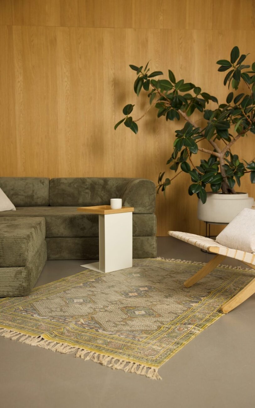 Ein modernes Wohnzimmer mit einem grünen Samtsofa, einem weißen Beistelltisch mit einer Tasse, einem gemusterten Teppich, einem hölzernen Akzentsessel mit einem weißen Kissen und einer großen Topfpflanze vor einer holzgetäfelten Wand.