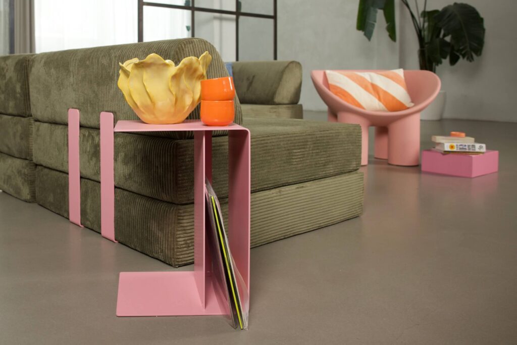 Ein modernes Wohnzimmer mit einem grünen Kordsofa, einem rosafarbenen Beistelltisch mit einer gelben Schale und einer orangefarbenen Tasse sowie einem rosafarbenen Stuhl mit einem gestreiften Kissen. Im Hintergrund sind eine Pflanze und Bücher zu sehen.