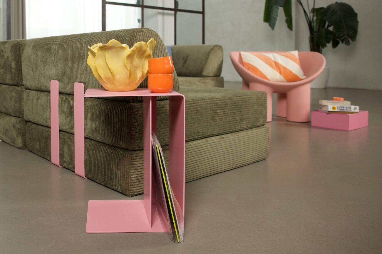 Ein modernes Wohnzimmer mit einem grünen Kordsofa, einem rosafarbenen Beistelltisch mit einer gelben Schale und einer orangefarbenen Tasse sowie einem rosafarbenen Stuhl mit einem gestreiften Kissen. Im Hintergrund sind eine Pflanze und Bücher zu sehen.