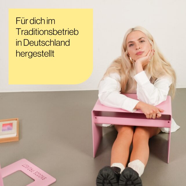 Eine Frau mit langen blonden Haaren sitzt auf dem Boden neben einem kleinen rosafarbenen Tisch und stützt ihren Kopf auf ihre Hand. Auf einem gelben Kasten mit deutschem Text steht: "Für dich im Traditionsbetrieb in Deutschland hergestellt.