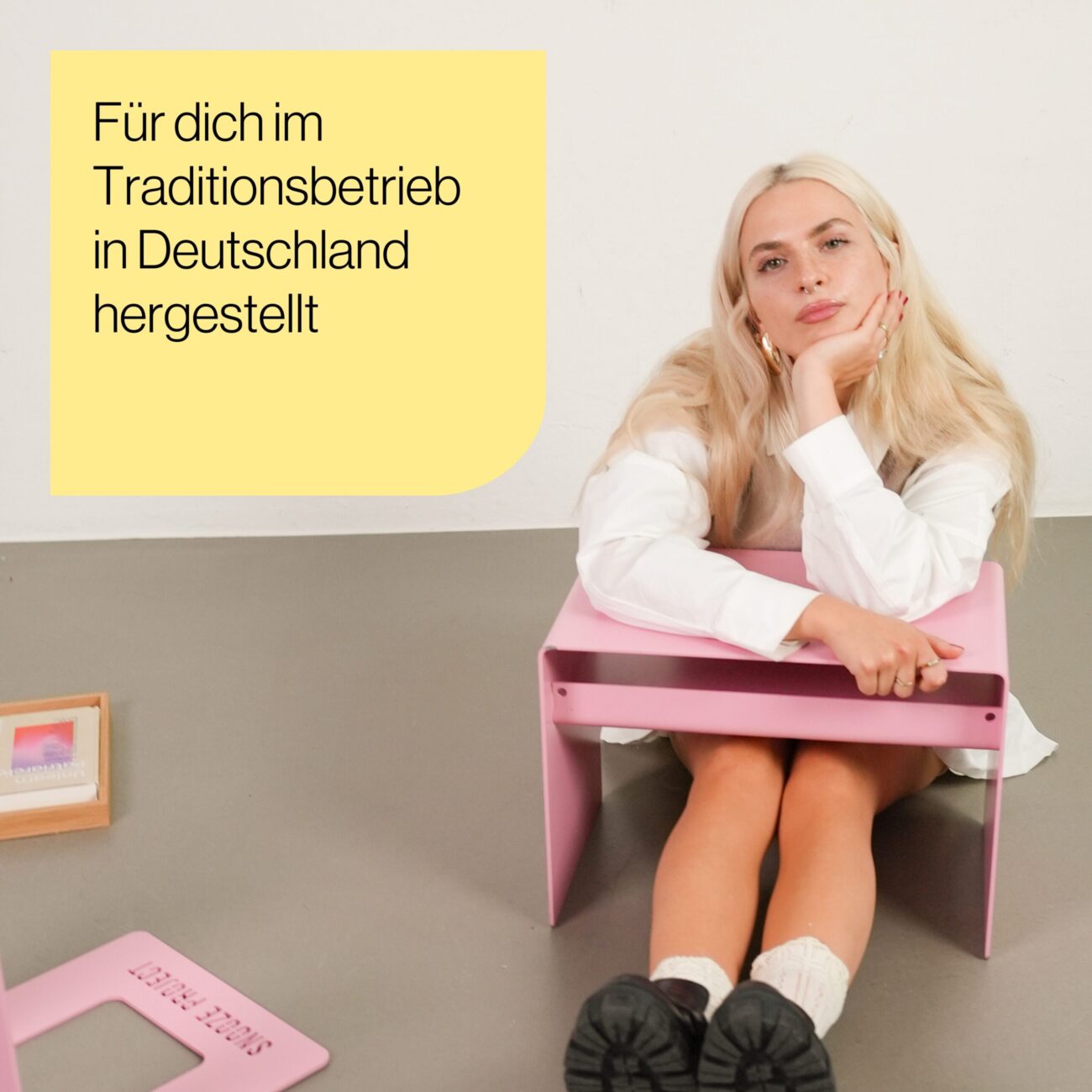 Tade_Produkt Mood_01 Eine Frau mit langen blonden Haaren sitzt auf dem Boden neben einem kleinen rosafarbenen Tisch und stützt ihren Kopf auf ihre Hand. Auf einem gelben Kasten mit deutschem Text steht: "Für dich im Traditionsbetrieb in Deutschland hergestellt.
