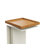 Ein quadratisches Holztablett mit erhöhten Rändern steht auf einem modernen weißen Sockel vor einem schlichten weißen Hintergrund.