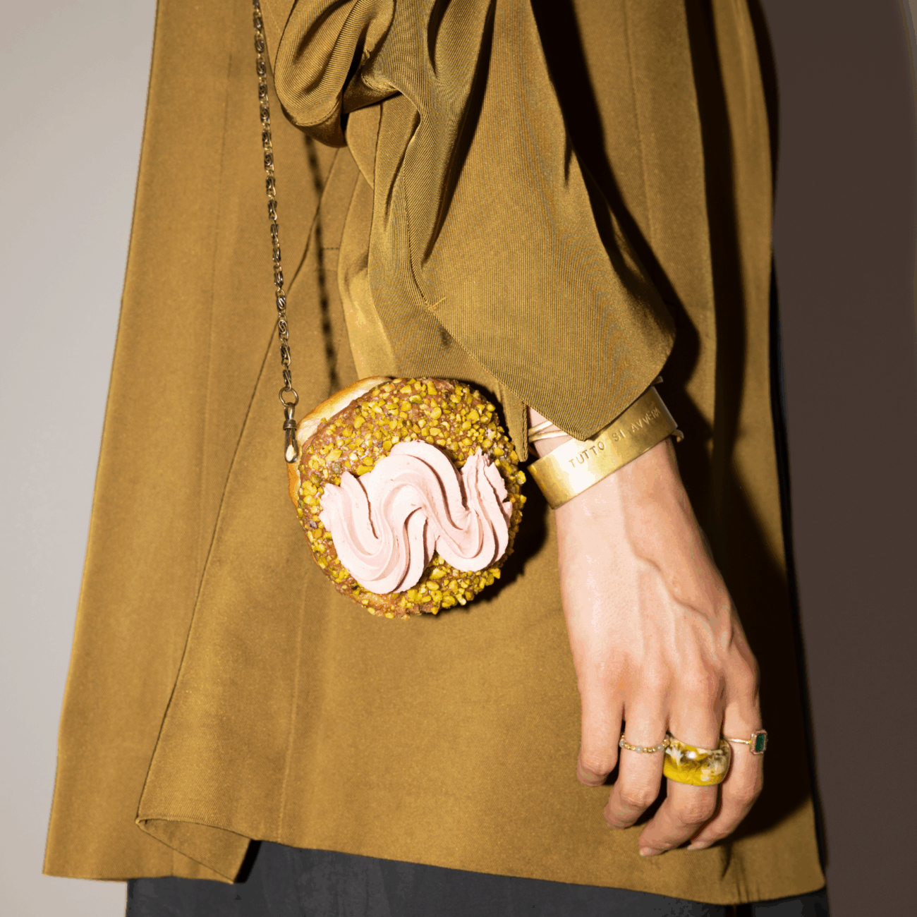 Eine Person, die einen braunen Mantel trägt, hält ein Portemonnaie in Form eines Donuts mit rosa Zuckerguss und gehackten Nüssen, das an einem Kettenband hängt. Die Person trägt goldene Ringe und einen Armreif.