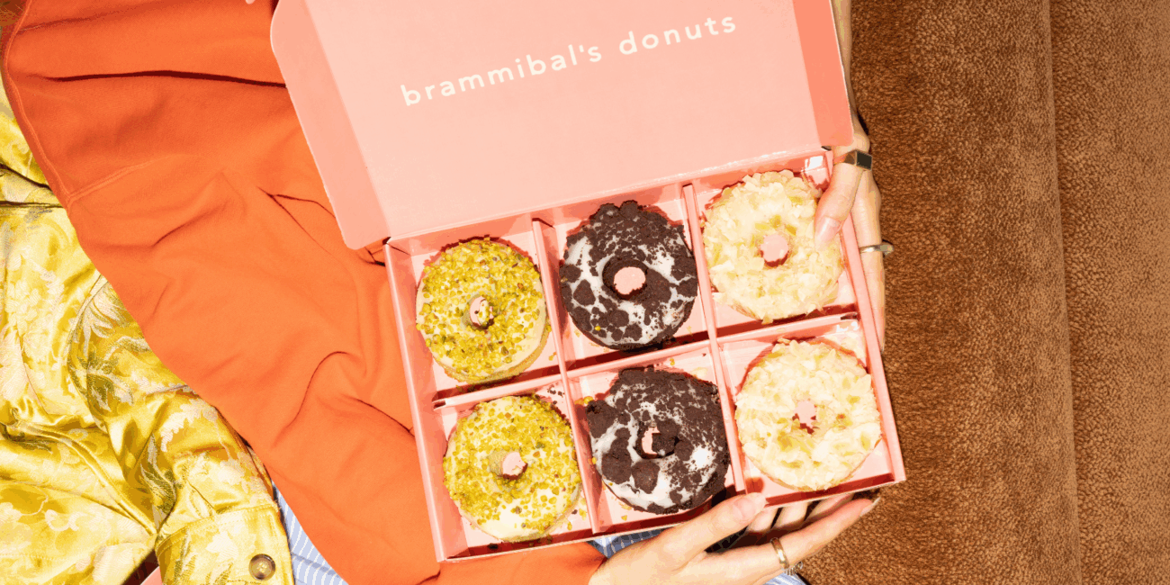 Eine Person hält eine geöffnete rosafarbene Schachtel mit der Aufschrift "Brammibals Donuts" in der Hand, die sechs verschiedene Donuts mit unterschiedlichen bunten Belägen enthält und auf einer braunen Fläche steht.