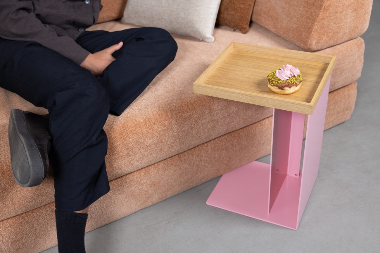 Eine Person sitzt auf einem pfirsichfarbenen Sofa neben einem kleinen Beistelltisch mit hölzerner Platte und rosafarbenem Sockel und hält einen Donut mit rosafarbener Glasur und zerstoßenen Nüssen in der Hand - bereit für ihren Hero-Moment im September 2025.