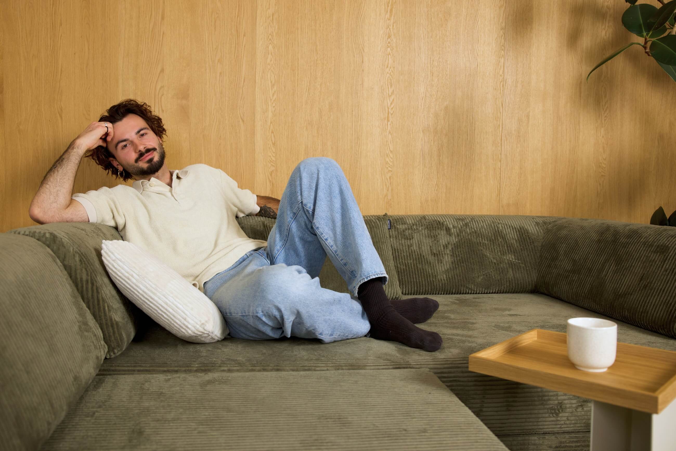 Ein Mann mit lockigem Haar und Bart sitzt auf einem grünen Sofa, das mit einem weißen Kissen abgestützt ist. Er trägt ein helles Hemd, blaue Jeans und schwarze Socken. Ein Kaffeebecher steht auf einem kleinen Holztisch in der Nähe.