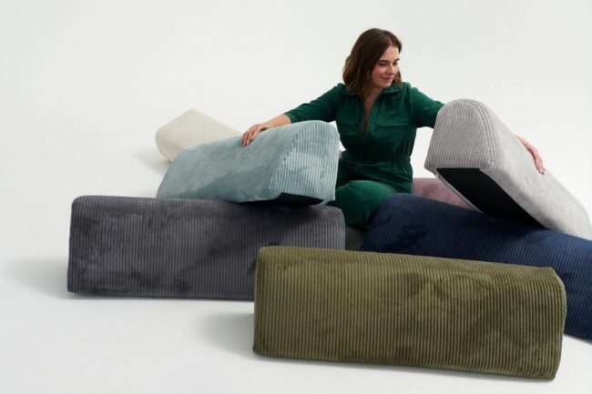 Eine Frau in grüner Kleidung sitzt auf dem Boden und arrangiert mehrere große, bunte Kordkissen in verschiedenen Formen und Farben auf weißem Hintergrund.