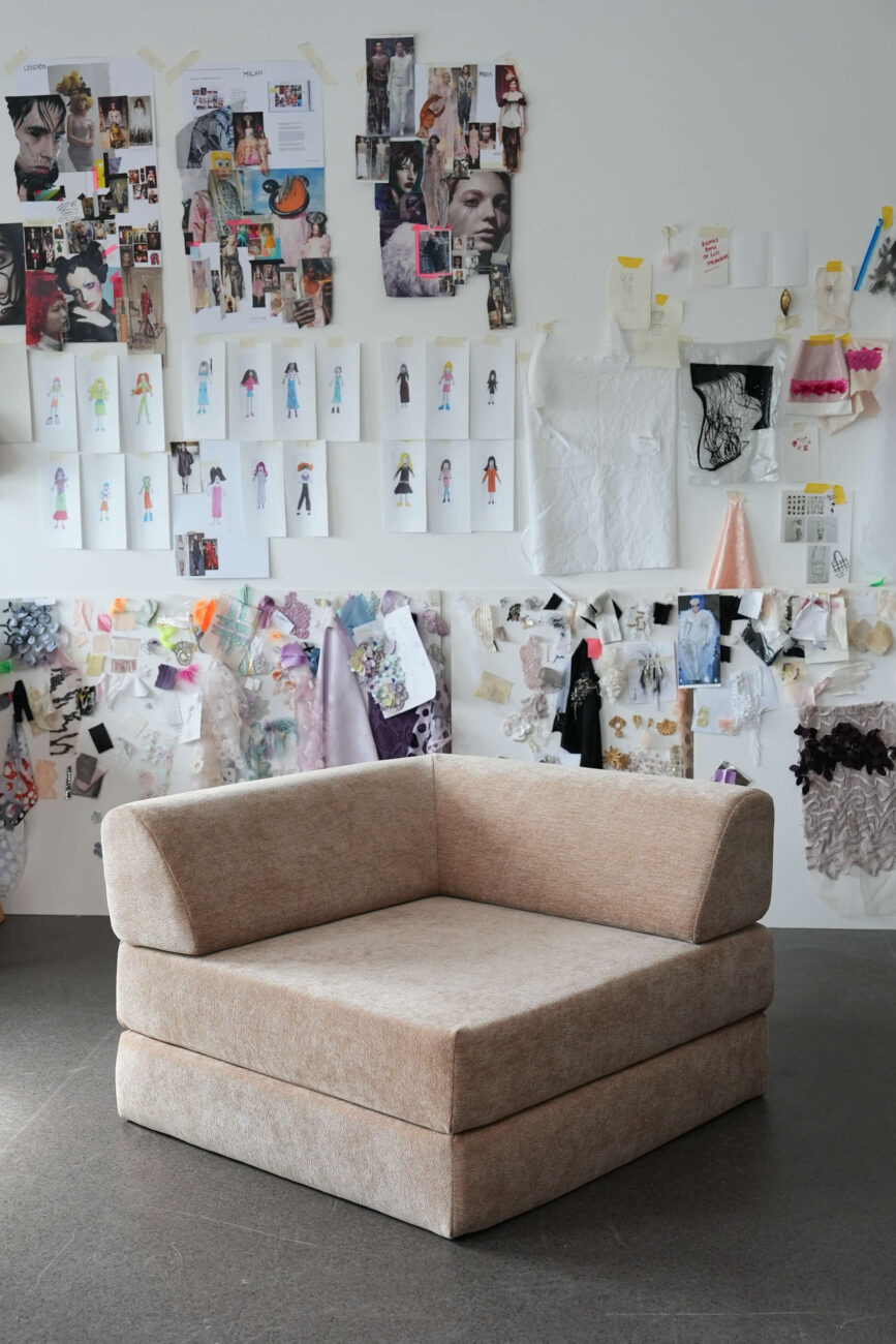 Ein weicher, beiger Ecksessel steht auf einem grauen Boden vor einer weißen Wand, die mit verschiedenen Modeskizzen, Stoffmustern, Zeitschriftenausschnitten und kreativen Designinspirationen bedeckt ist.