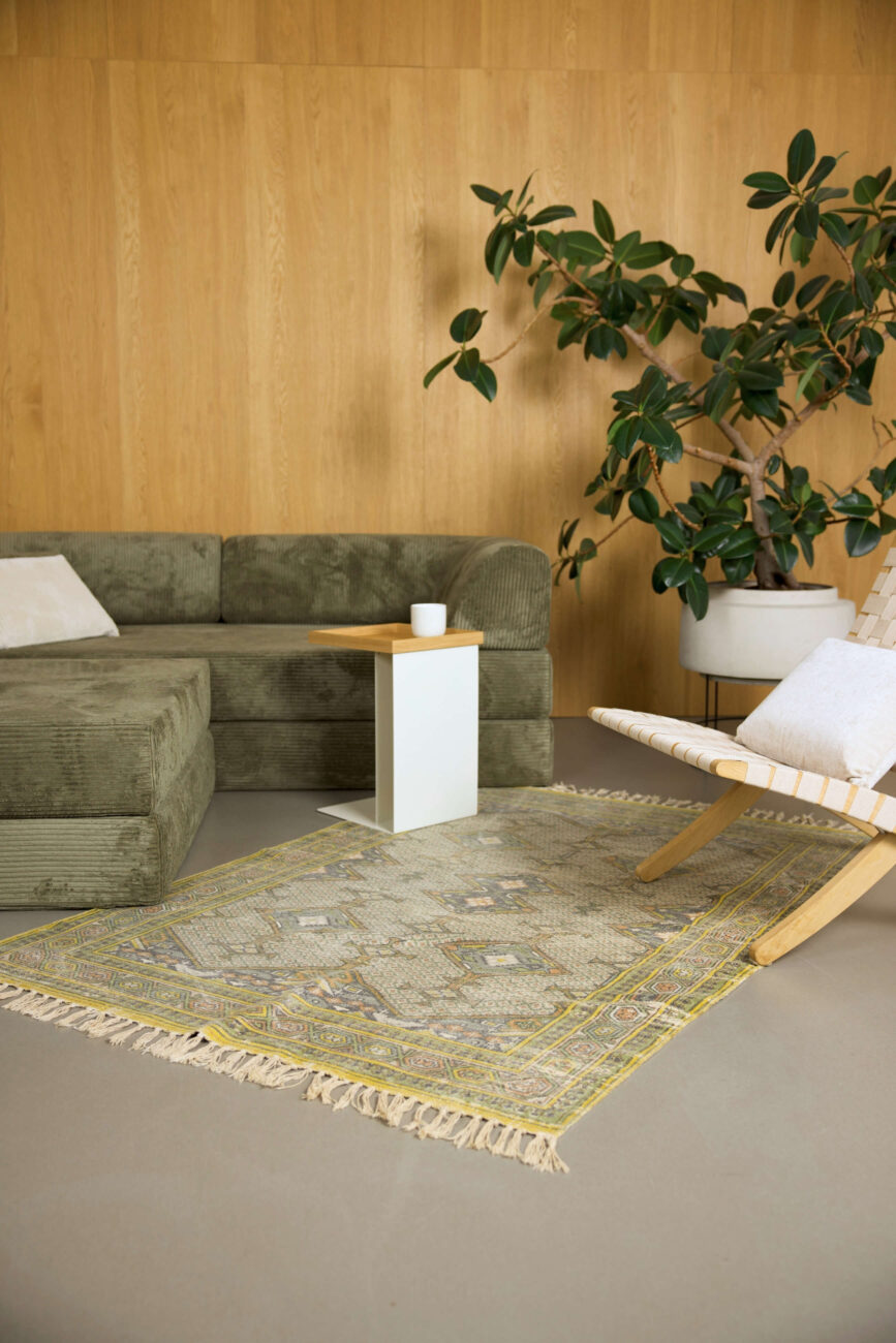 Ein modernes Wohnzimmer mit einem grünen Sektionssofa, einem kleinen weißen Beistelltisch mit einer Tasse, einem gemusterten Teppich, einem Holzstuhl mit einem Kissen und einer großen Topfpflanze vor einer Holzwand.