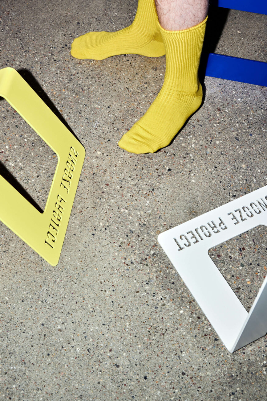 Eine Person, die leuchtend gelbe Socken trägt, steht auf einem gesprenkelten Boden in der Nähe von zwei metallenen Buchstützen mit der Aufschrift PROJECT PROJECT in Gelb und Weiß, mit einem blauen Objekt im Hintergrund.