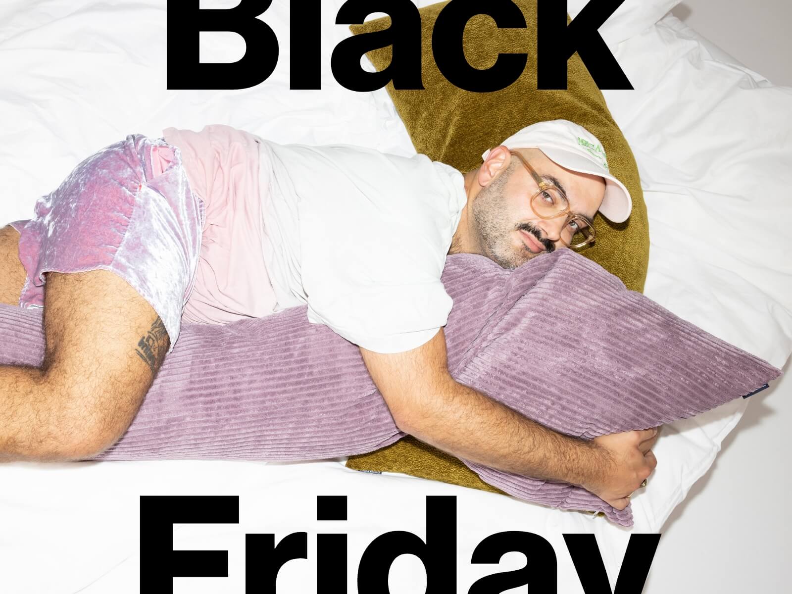 Ein Mann in pastellfarbenen Shorts, einem weißen T-Shirt und einer Mütze liegt auf einem Bett und umarmt ein lila Kissen. Über ihm steht in großer schwarzer Schrift "Black Friday".