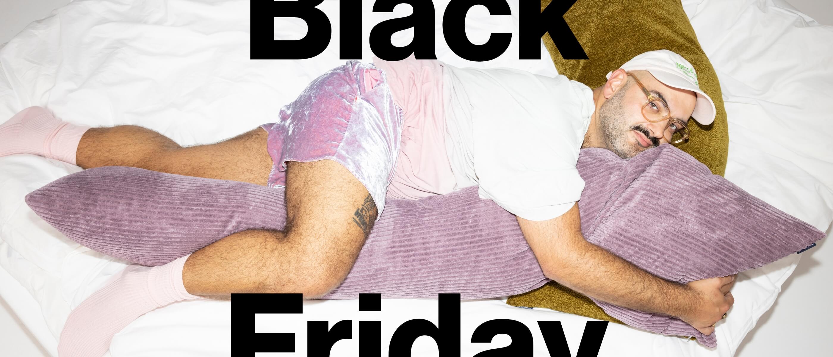 Ein Mann in rosa Shorts, einem weißen T-Shirt und einer Mütze umarmt ein langes lila Kissen, während er auf einem Bett liegt. Die Worte Black Friday stehen in großer schwarzer Schrift über dem Bild.
