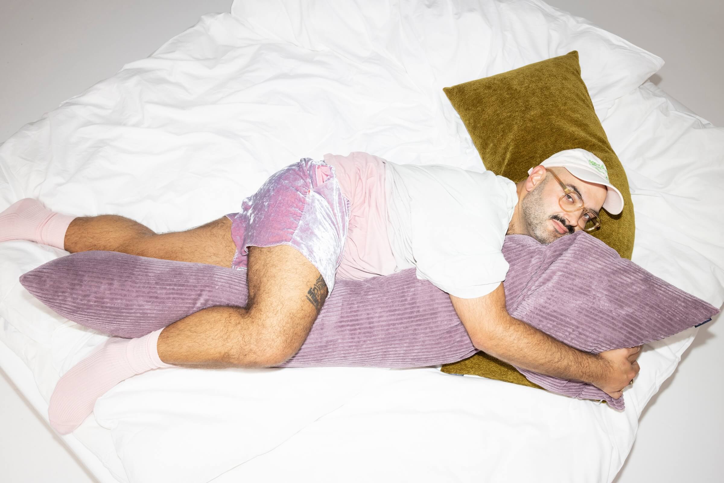 Ein Mann, der pastellfarbene Kleidung, rosa Socken und eine Mütze trägt, liegt auf einem Bett, umarmt ein großes lila Kissen und ruht sich auf einem olivgrünen Kissen aus, wobei er entspannt und bequem aussieht.