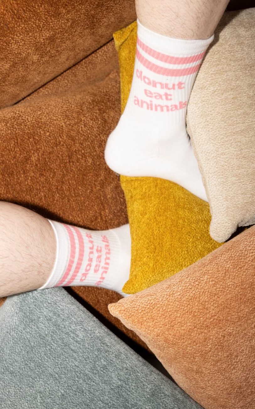 Eine Person, die weiße Socken mit rosa Streifen und der Aufschrift "Donut eat animals" trägt, legt ihre Füße auf einen Stapel bunter, strukturierter Kissen.