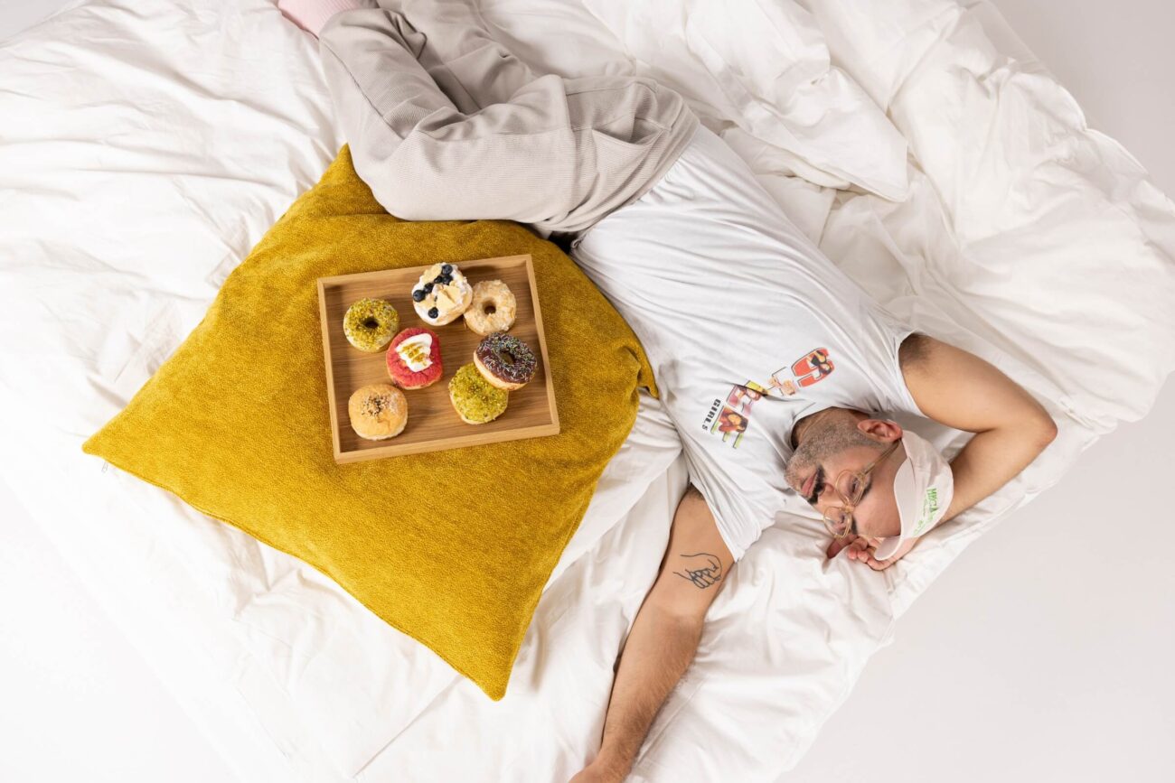 Eine Person mit Schlafmaske und Freizeitkleidung liegt auf einem Bett neben einem gelben Kissen mit einem Holztablett mit verschiedenen Donuts.