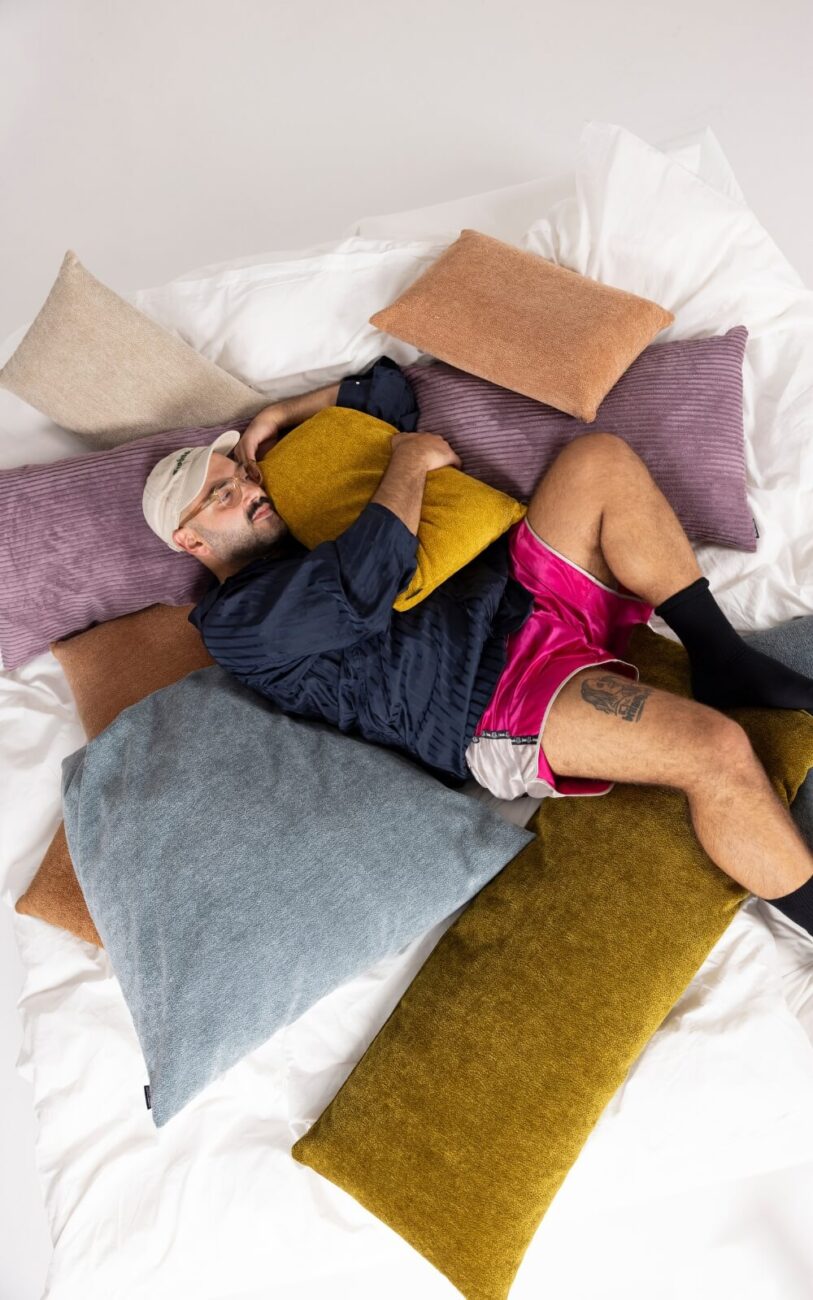 Eine Person, die eine Mütze, ein dunkles Hemd und hellrosa Shorts trägt, liegt auf einem mit bunten Kissen bedeckten Bett, umarmt ein gelbes Kissen, hat die Beine gekreuzt und sieht entspannt aus.