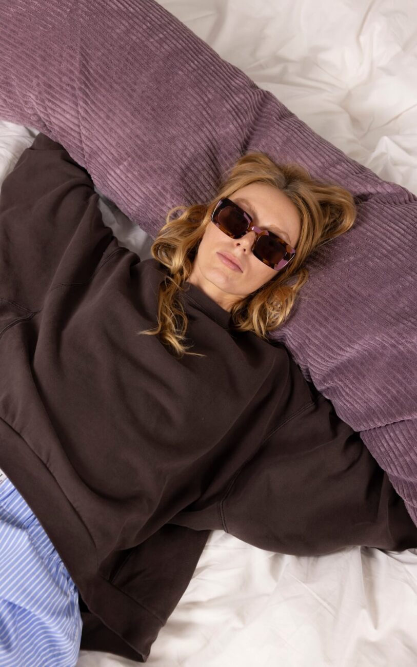 Eine Frau mit Sonnenbrille und dunklem Sweatshirt liegt auf einem Bett mit einem lila Kissen hinter dem Kopf und sieht entspannt aus. Außerdem trägt sie eine hellblau gestreifte Hose.