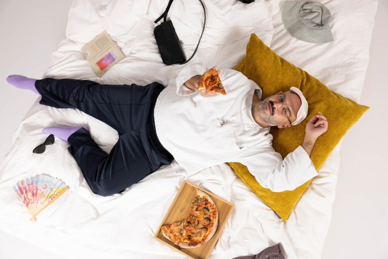 Ein Mann liegt auf einem Bett mit einem gelben Kissen und hält ein Stück Pizza in der Hand. Ein Tablett mit noch mehr Pizza, ein Buch, ein Hut, eine Handtasche, eine Sonnenbrille, ein Fächer und ein Hemd sind um ihn herum auf dem weißen Bettzeug verstreut.