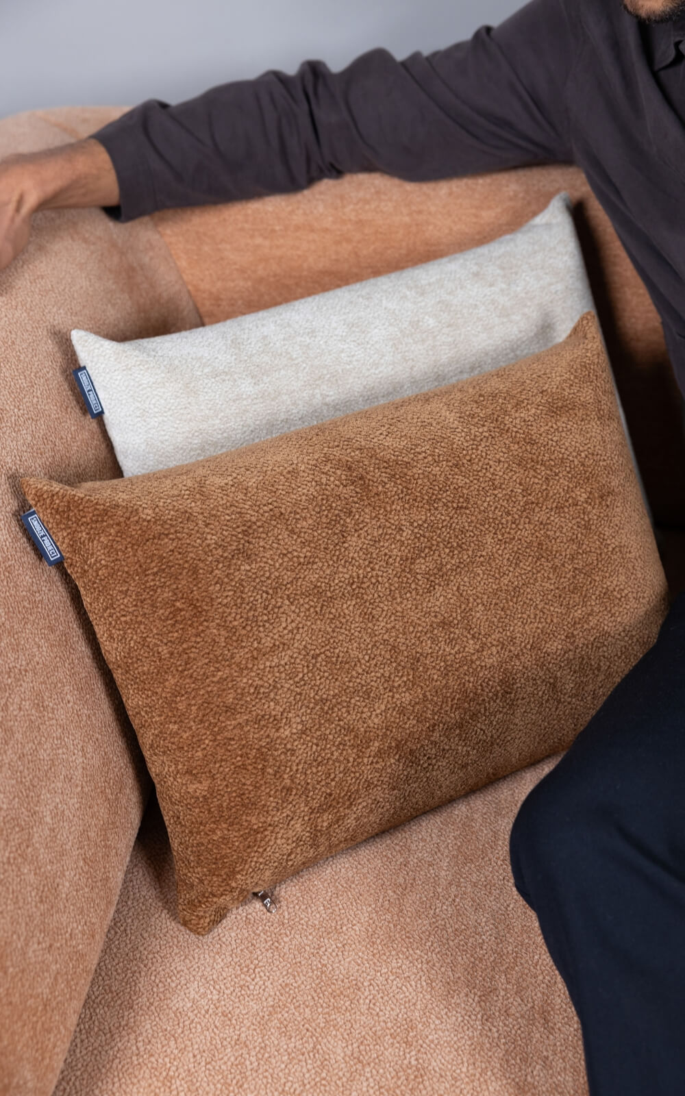Eine Person sitzt auf einer beigen Couch mit zwei rechteckigen Kissen, einem hellbeigen und einem braunen, die auf dem Sitz ruhen. Man sieht den Arm der Person, die mit einem langärmeligen dunklen Hemd bekleidet ist.