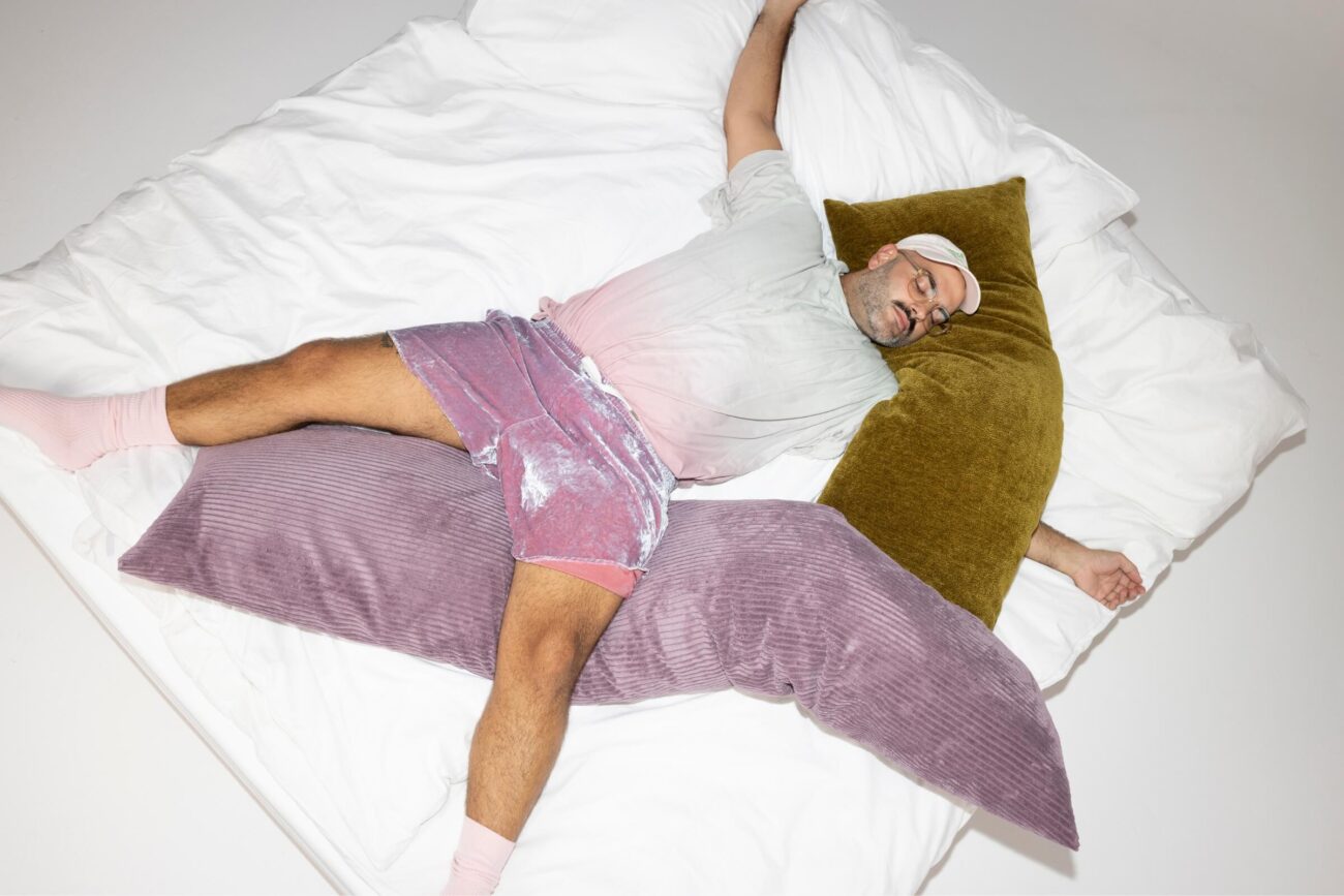 Eine Person, die eine Schlafmaske, ein T-Shirt, Shorts und Socken trägt, liegt auf einem Bett mit weißen Laken und ruht auf einem großen olivgrünen Kissen und einem großen lavendelfarbenen Kissen. Die Szene ist hell beleuchtet.