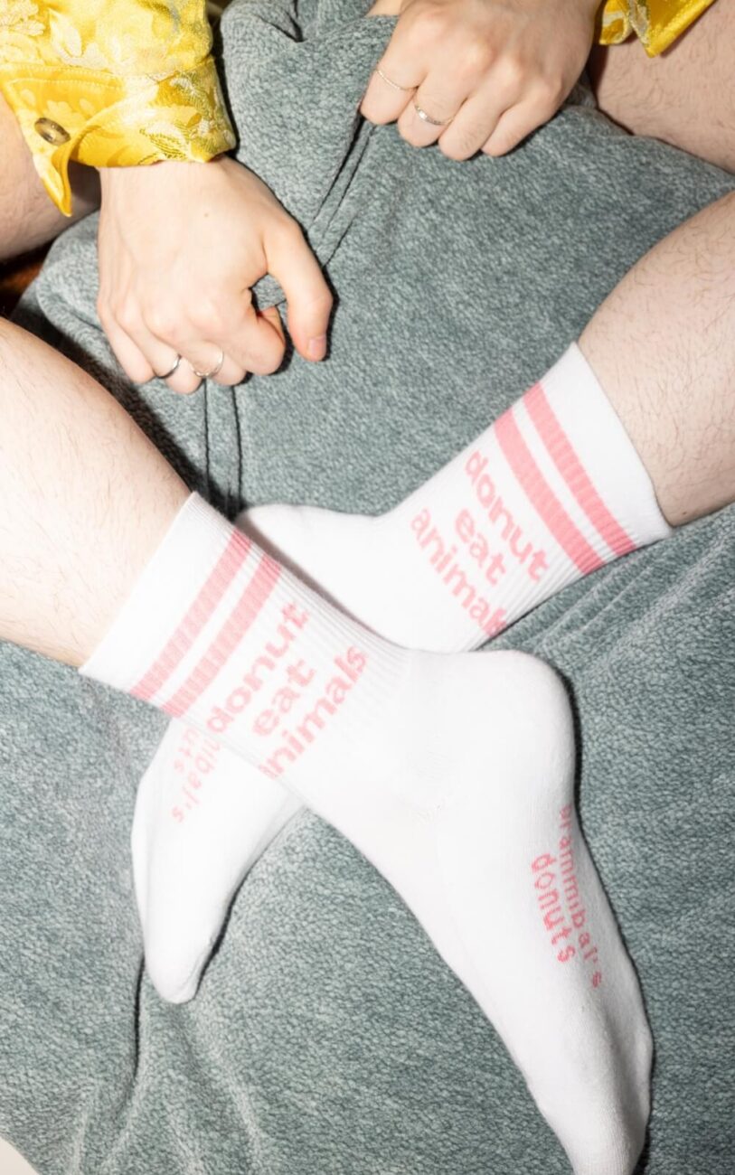 Eine Person, die weiße Socken mit rosa Streifen und der Aufschrift "Donut eat animals" trägt, sitzt mit gekreuzten Beinen auf einer grünen Decke. Ihre Hände liegen auf dem Schoß, und ein Teil eines gelben Kleidungsstücks ist zu sehen.
