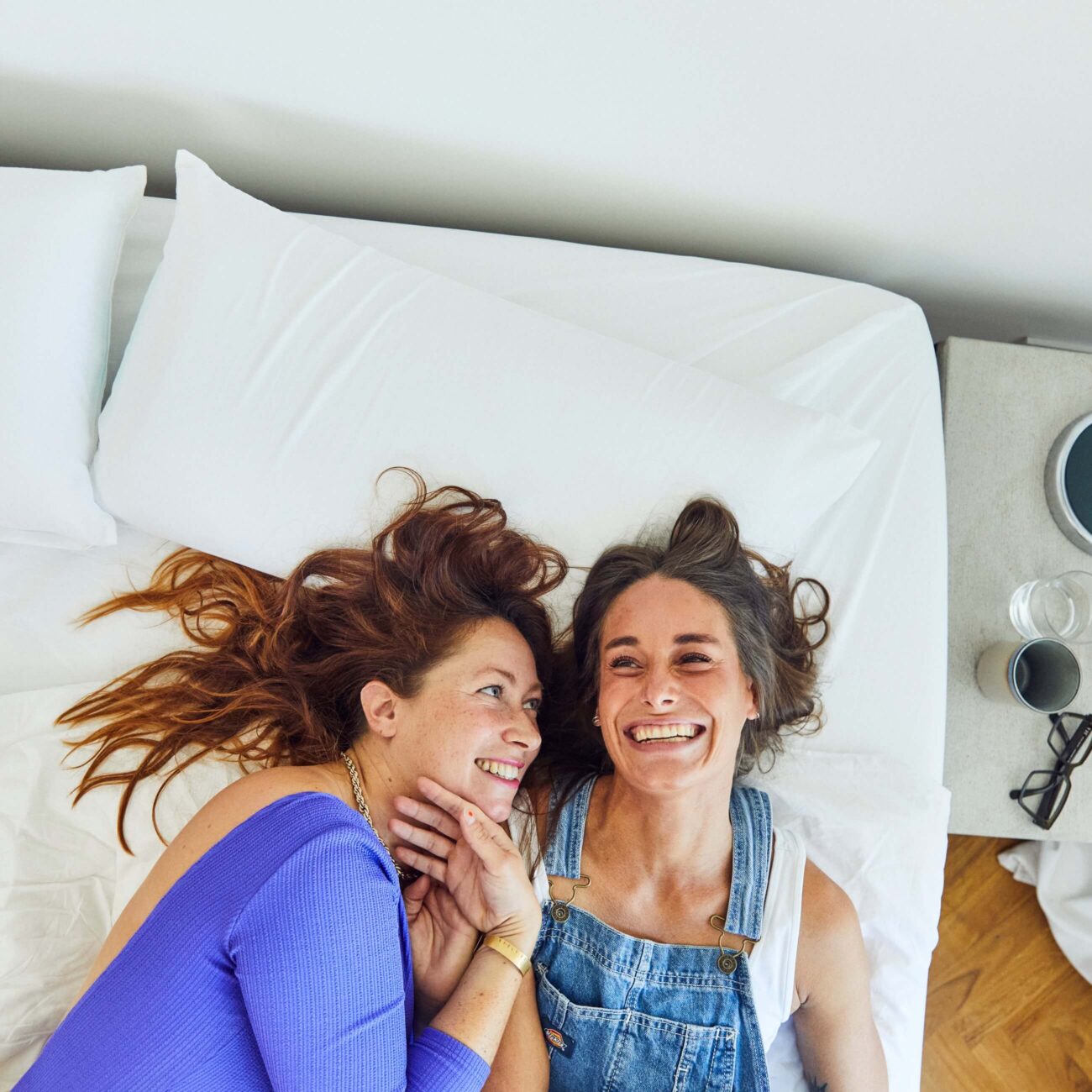 SP-Mood_img001 Zwei Frauen liegen auf einem Bett, lächeln sich an und schauen sich an. Die eine trägt ein blaues Oberteil, die andere einen Jeans-Overall. Sie wirken glücklich und entspannt, ihre Köpfe liegen eng beieinander auf dem weißen Bettzeug.