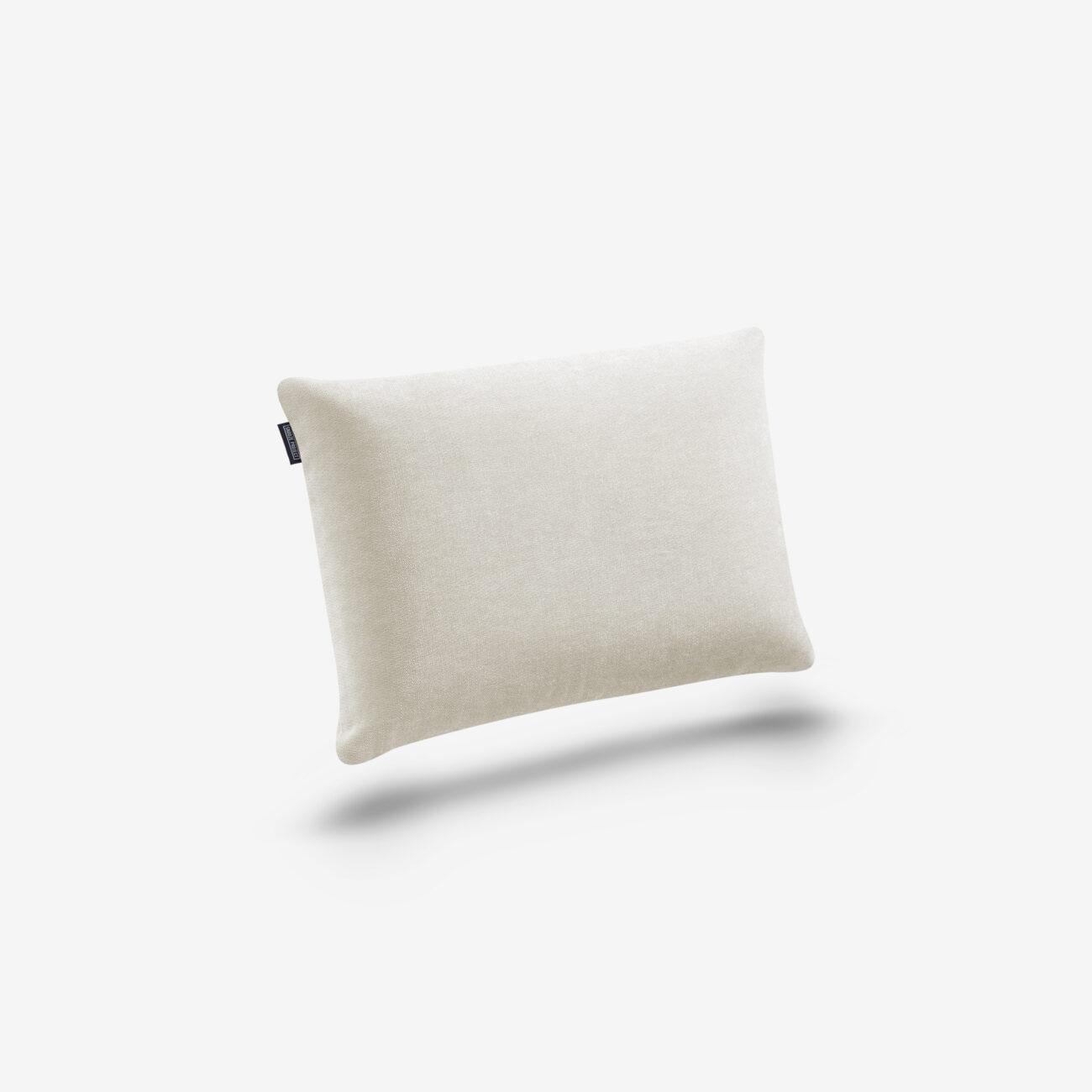 SPKDEKO45x30-SC-TW-img001 Ein rechteckiges, cremefarbenes Kissen mit einem kleinen dunklen Etikett an der Seite, das sich vor einem weißen Hintergrund mit einem dezenten Schatten darunter befindet.