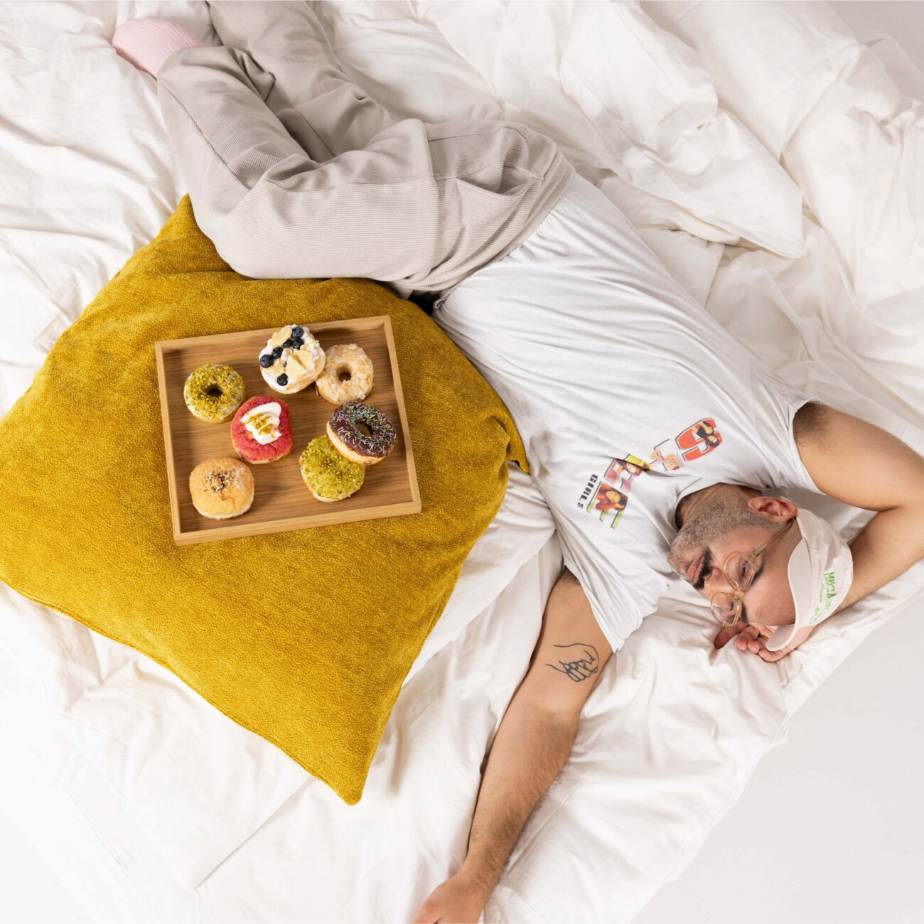 SPKOTTI80x80-Mood-img001 Ein Mann liegt auf einem weißen Bett mit einem gelben Kissen neben sich und hält ein Holztablett mit verschiedenen verzierten Donuts in der Hand. Er trägt eine Brille, ein Stirnband, ein weißes T-Shirt und eine hellgraue Hose.