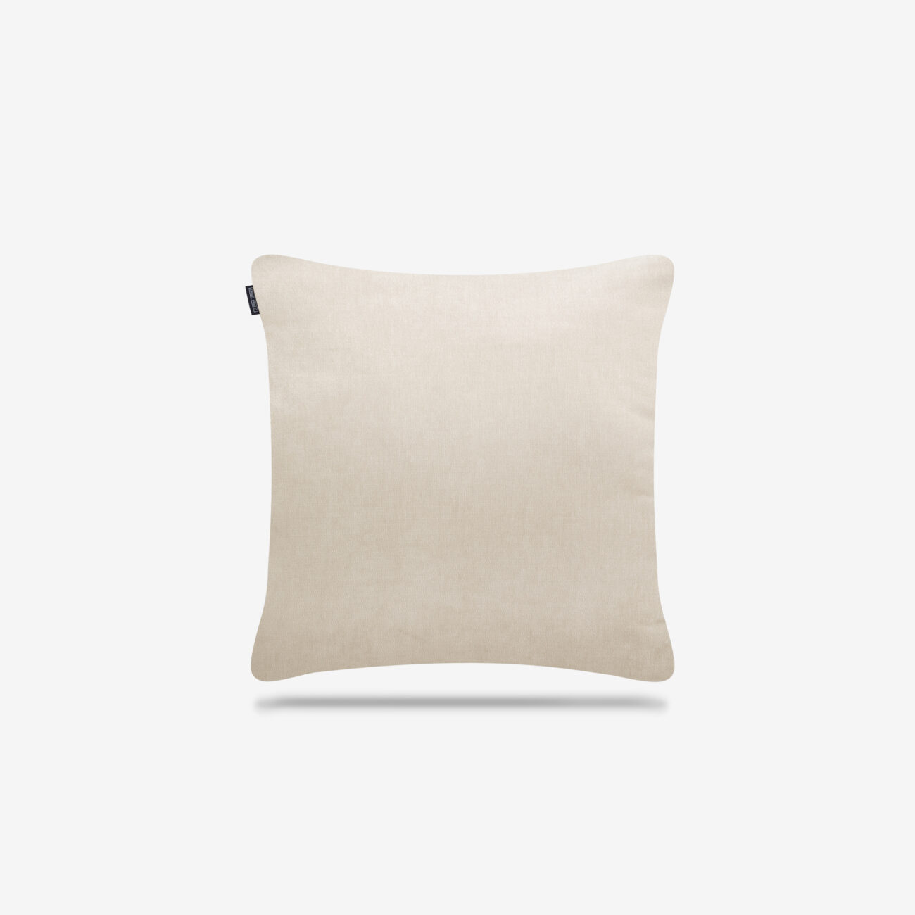 Ein schlichtes, quadratisches, beigefarbenes Kissen mit einem kleinen schwarzen Etikett auf der linken Seite, das sich vor einem weißen Hintergrund mit einem schwachen Schatten darunter befindet.