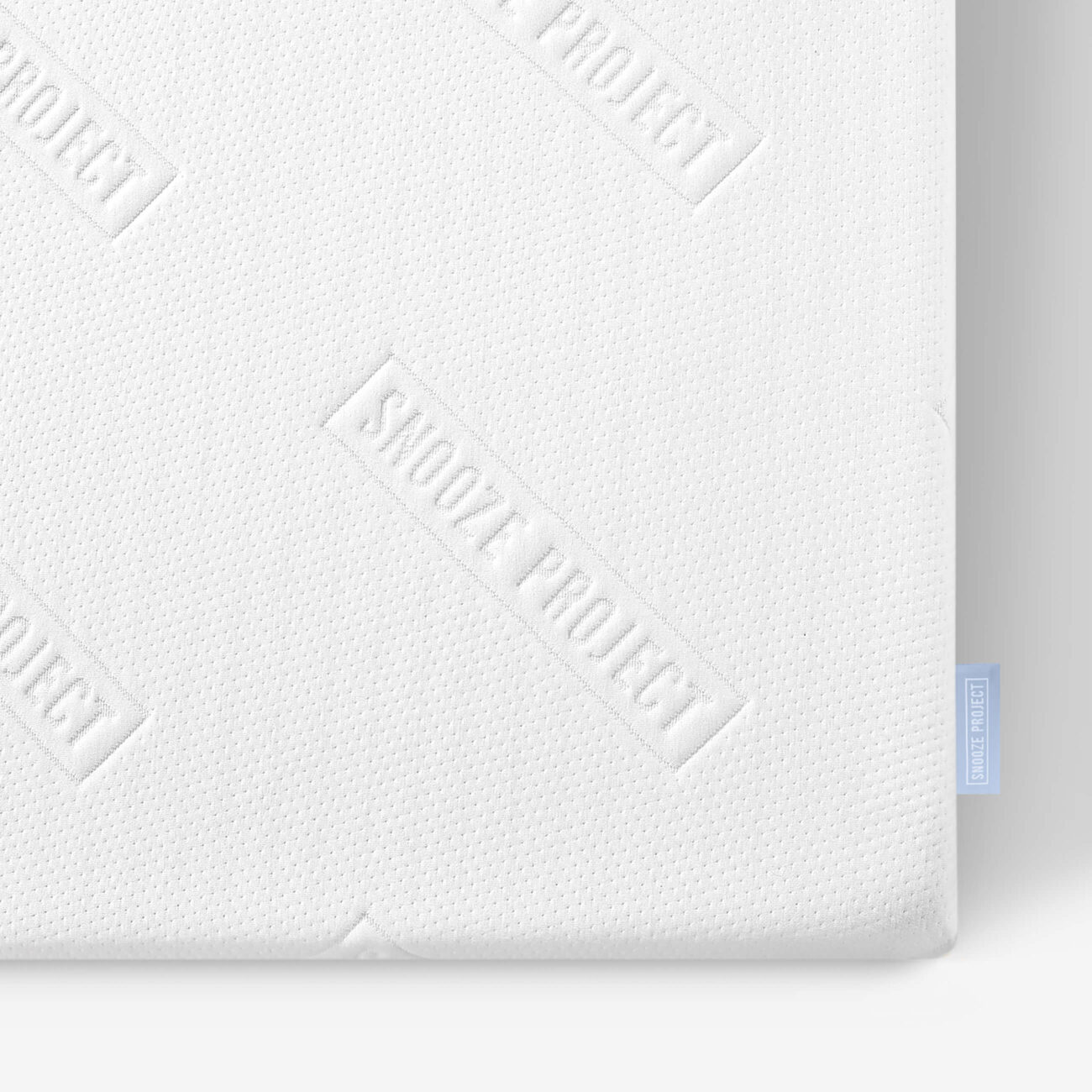 SPS_img004 Nahaufnahme einer weißen Matratze mit dezent gepunkteter Struktur und dem wiederholt auf die Oberfläche geprägten Schriftzug SKOOZE PROJECT; am Rand der Matratze ist ein kleines blaues Stoffschild angebracht.