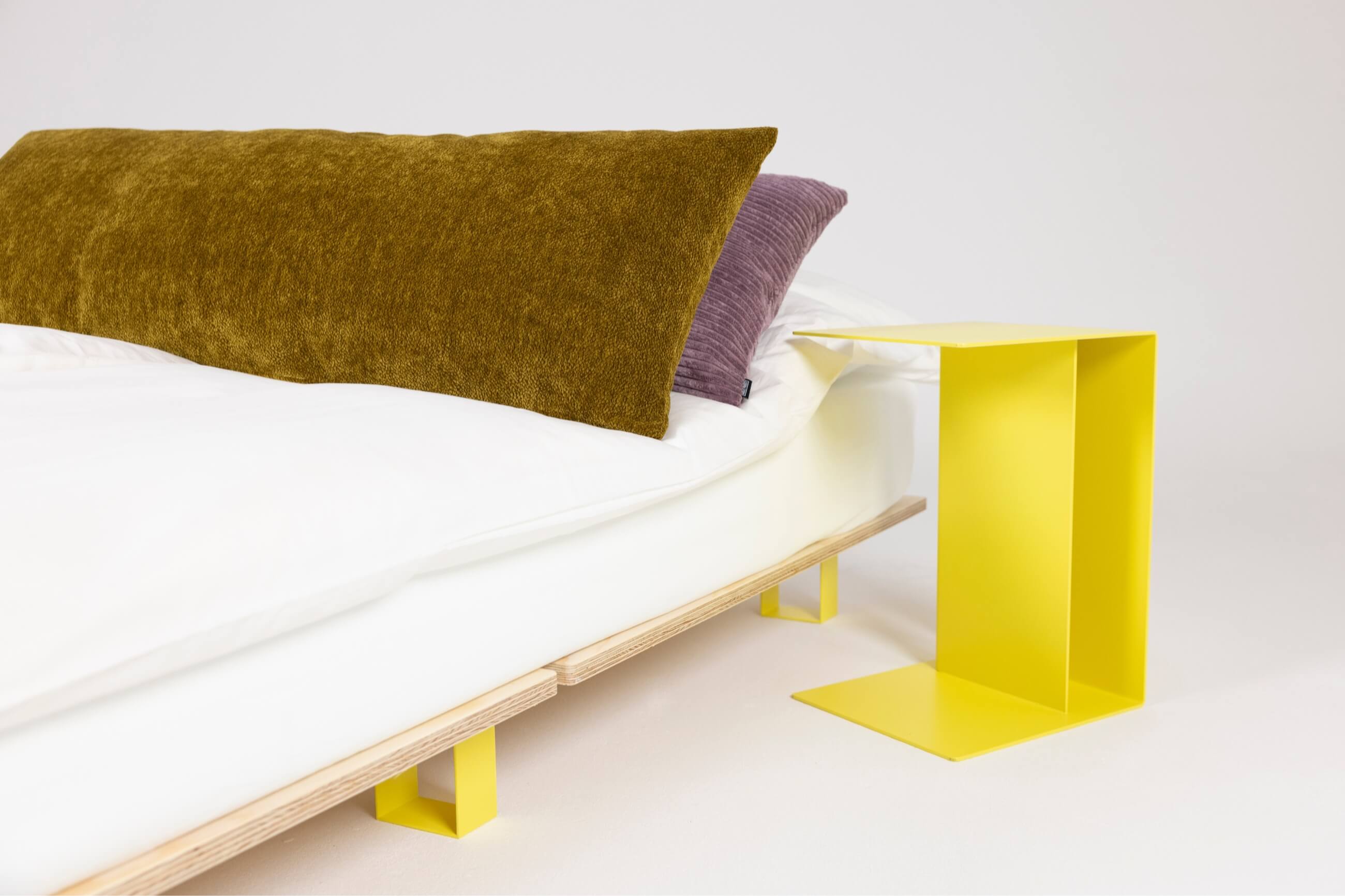 Ein modernes Bett mit weißem Bettzeug, einem braunen rechteckigen Kissen und einem lila Kissen. Neben dem Bett steht ein minimalistischer, leuchtend gelber Beistelltisch aus Metall mit geometrischem Muster.