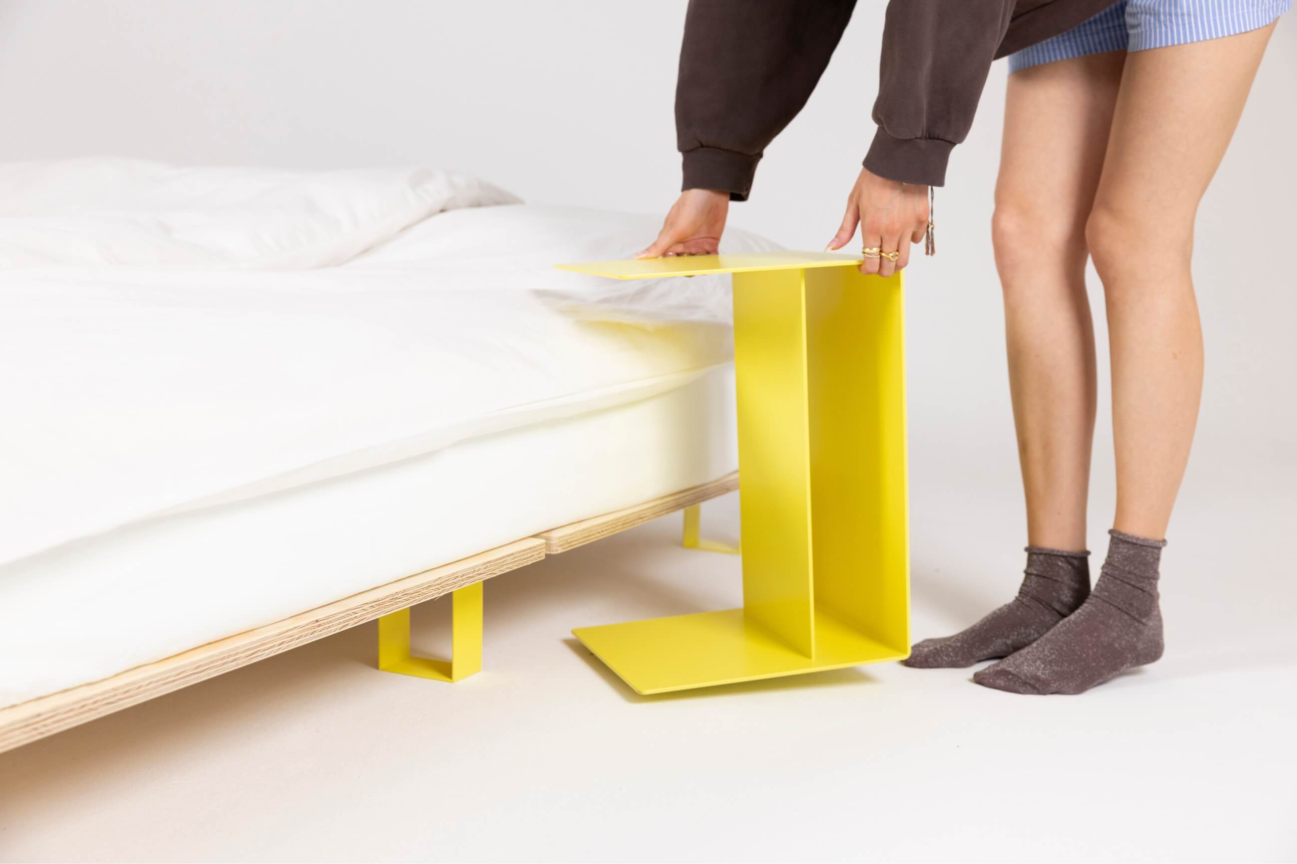 Eine Person stellt einen hellgelben, L-förmigen Beistelltisch neben ein niedriges, modernes Bett mit weißem Bettzeug. Die Person trägt einen dunklen Pullover, blaue Shorts und graue Socken. Der Hintergrund ist einfarbig und hell.