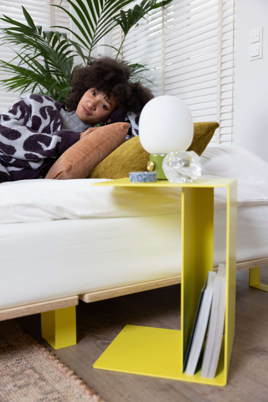Eine Frau mit lockigem Haar liegt auf einem Bett mit senfgelben und braunen Kissen, zugedeckt von einer gemusterten Decke. Neben ihr steht ein moderner gelber Nachttisch mit einer Lampe, Kristall und Zeitschriften. Große Grünpflanze im Hintergrund.