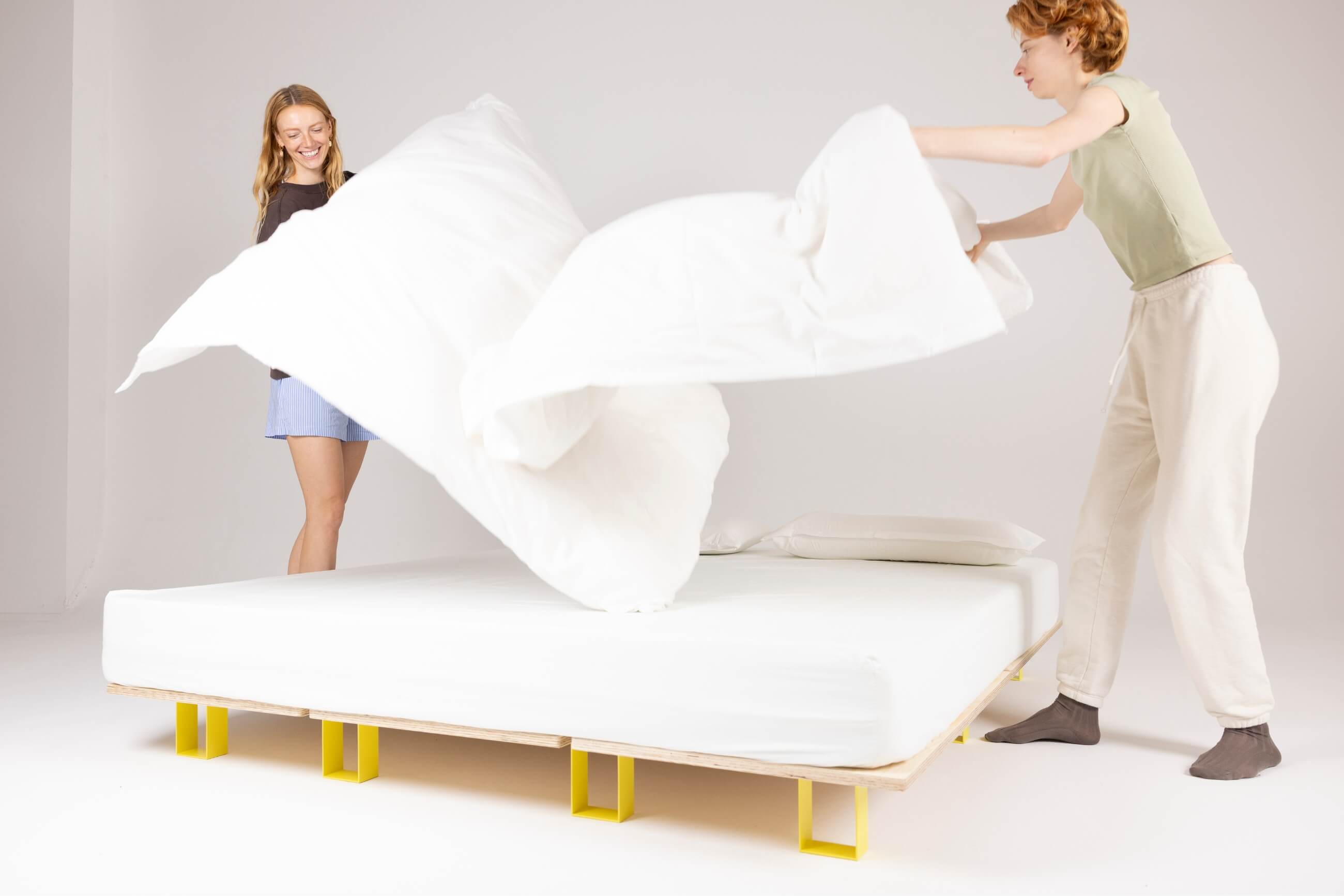 Zwei Menschen lächeln und machen gemeinsam ein Bett, indem sie eine weiße Bettdecke über eine Matratze auf einem modernen, niedrigen Bettgestell mit gelben Beinen in einem hellen, minimalistischen Raum heben.