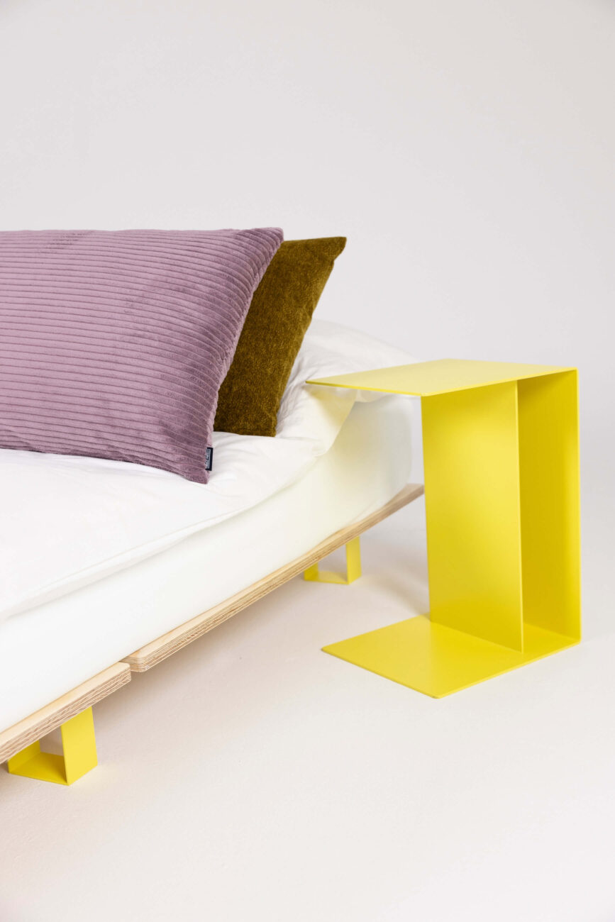 Ein modernes Bett mit weißer Matratze und zwei Kissen - eines lavendelfarben, eines olivfarben - neben einem minimalistischen, leuchtend gelben Beistelltisch aus Metall mit einem geometrischen Muster auf weißem Hintergrund.