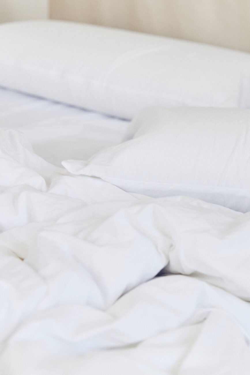 Nahaufnahme eines ungemachten Bettes mit weißen Laken und zwei weißen Kissen, von denen eines flach liegt und das andere leicht angewinkelt ist. Das Bettzeug wirkt weich und einladend, mit sanften Falten und Knicken.