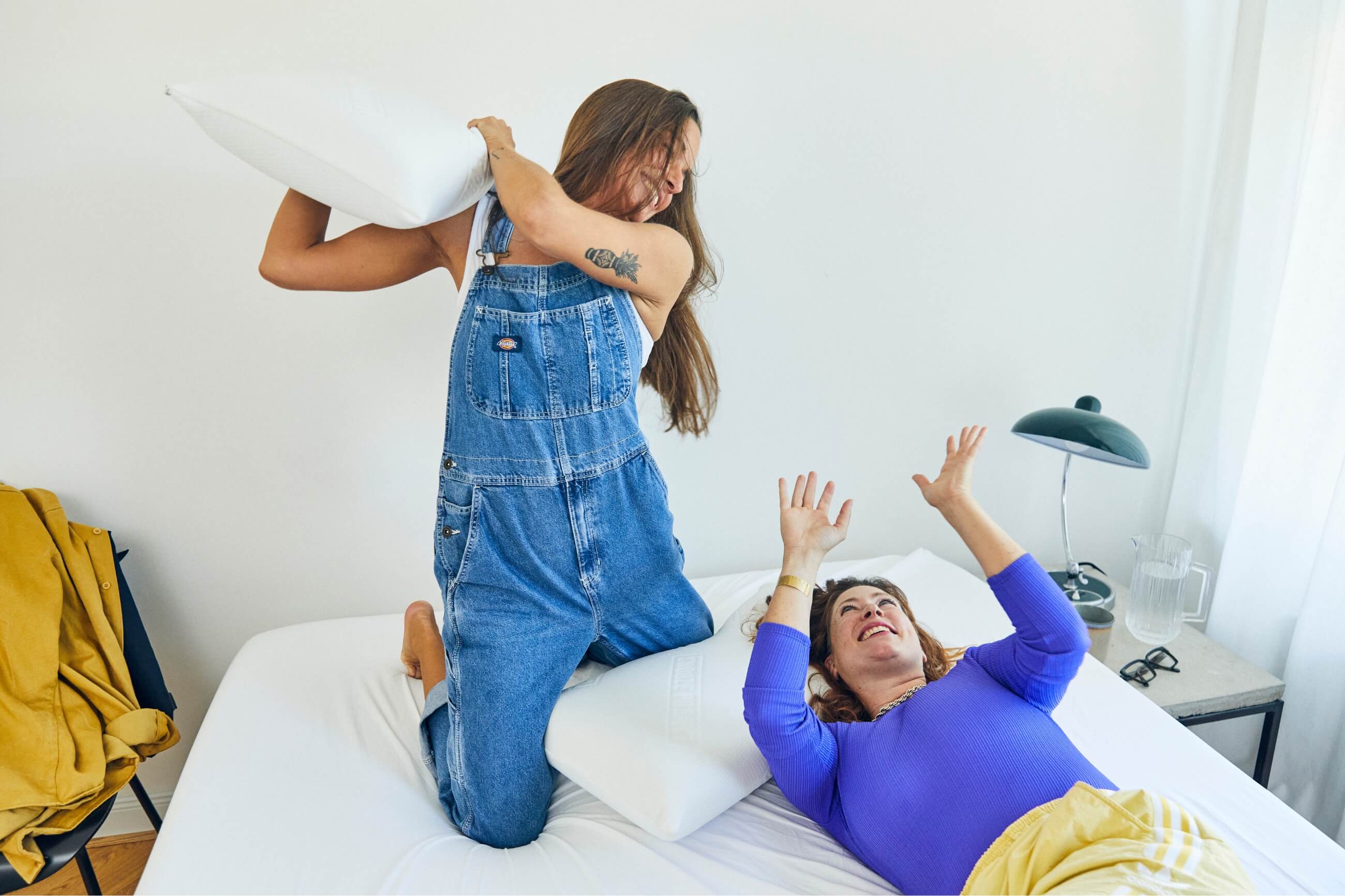 Zwei Frauen liefern sich auf einem Bett eine spielerische Kissenschlacht. Eine Frau kniet und hält ein Kissen über ihren Kopf, während die andere liegt, lächelt und ihre Hände zur Verteidigung hebt.