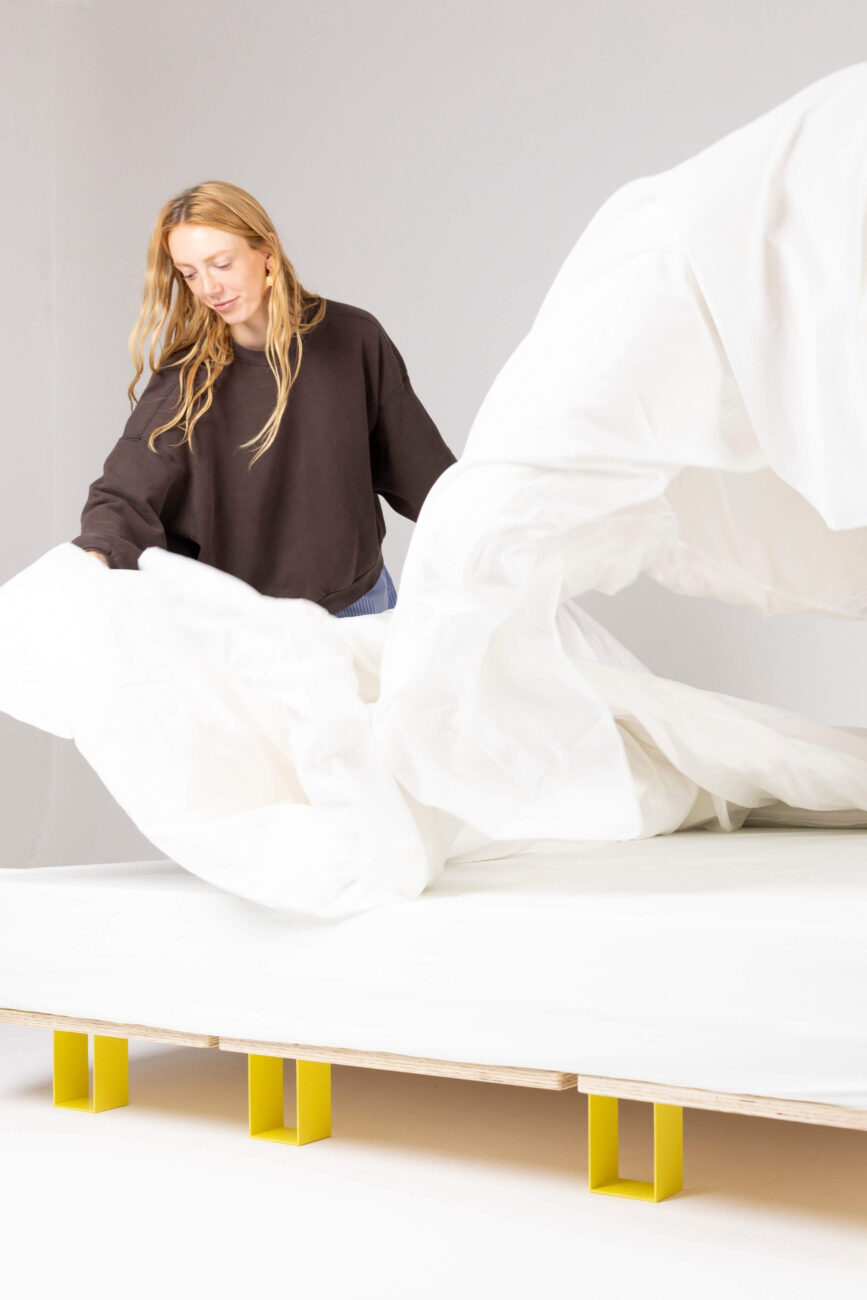 Eine Frau mit langen blonden Haaren, die ein schwarzes Sweatshirt und eine blaue Hose trägt, breitet ein großes weißes Laken über ein Bett mit einem einfachen, modernen Rahmen und gelben Beinen aus.