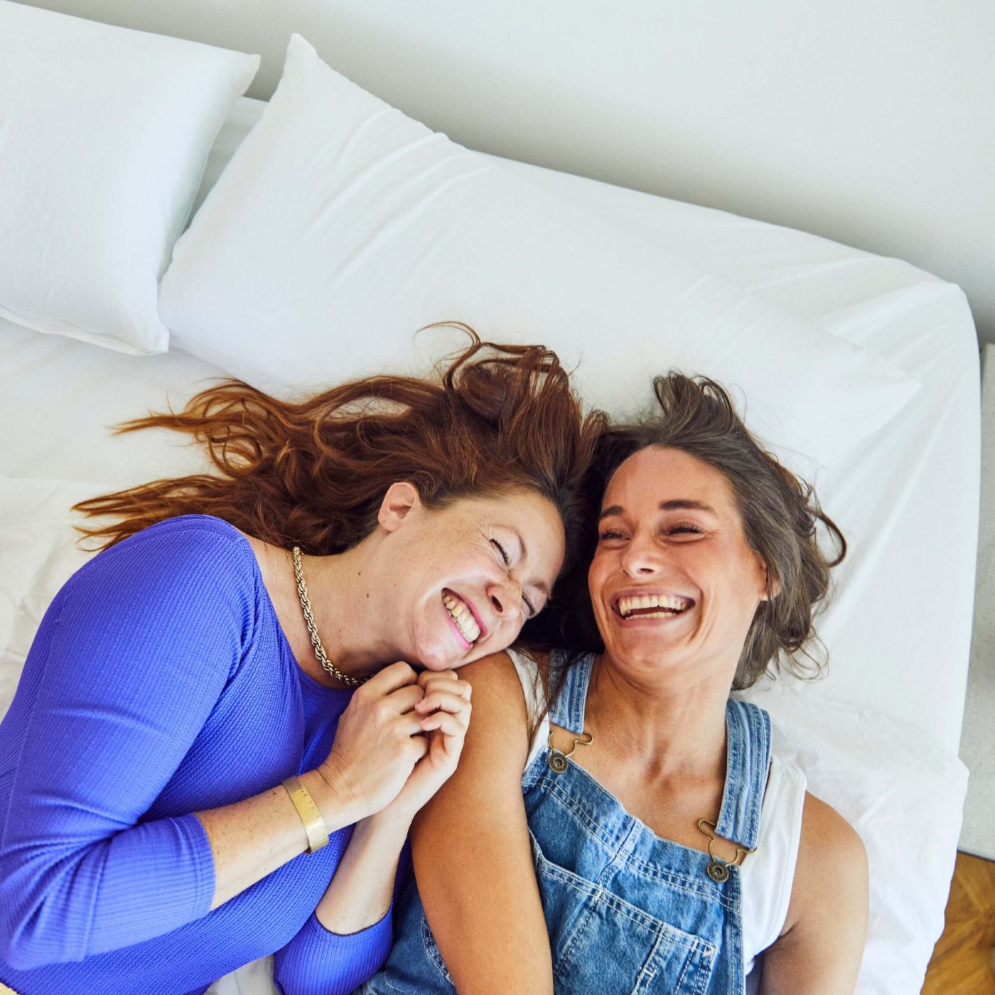 SPTUVAUSSM-Mood_img001 Zwei Frauen liegen auf einem Bett, lächeln und lachen zusammen. Die eine trägt ein blaues langärmeliges Oberteil, die andere einen Jeans-Overall über einem weißen Hemd. Sie sehen glücklich und entspannt aus und genießen die Gesellschaft des jeweils anderen.