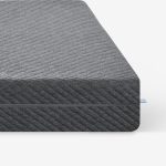 Nahaufnahme der Ecke einer dunkelgrauen, strukturierten Matratze mit einem dezenten diagonalen Streifenmuster vor einem schlichten weißen Hintergrund. Ein kleines blaues Stoffetikett ist an der Matratzennaht sichtbar.