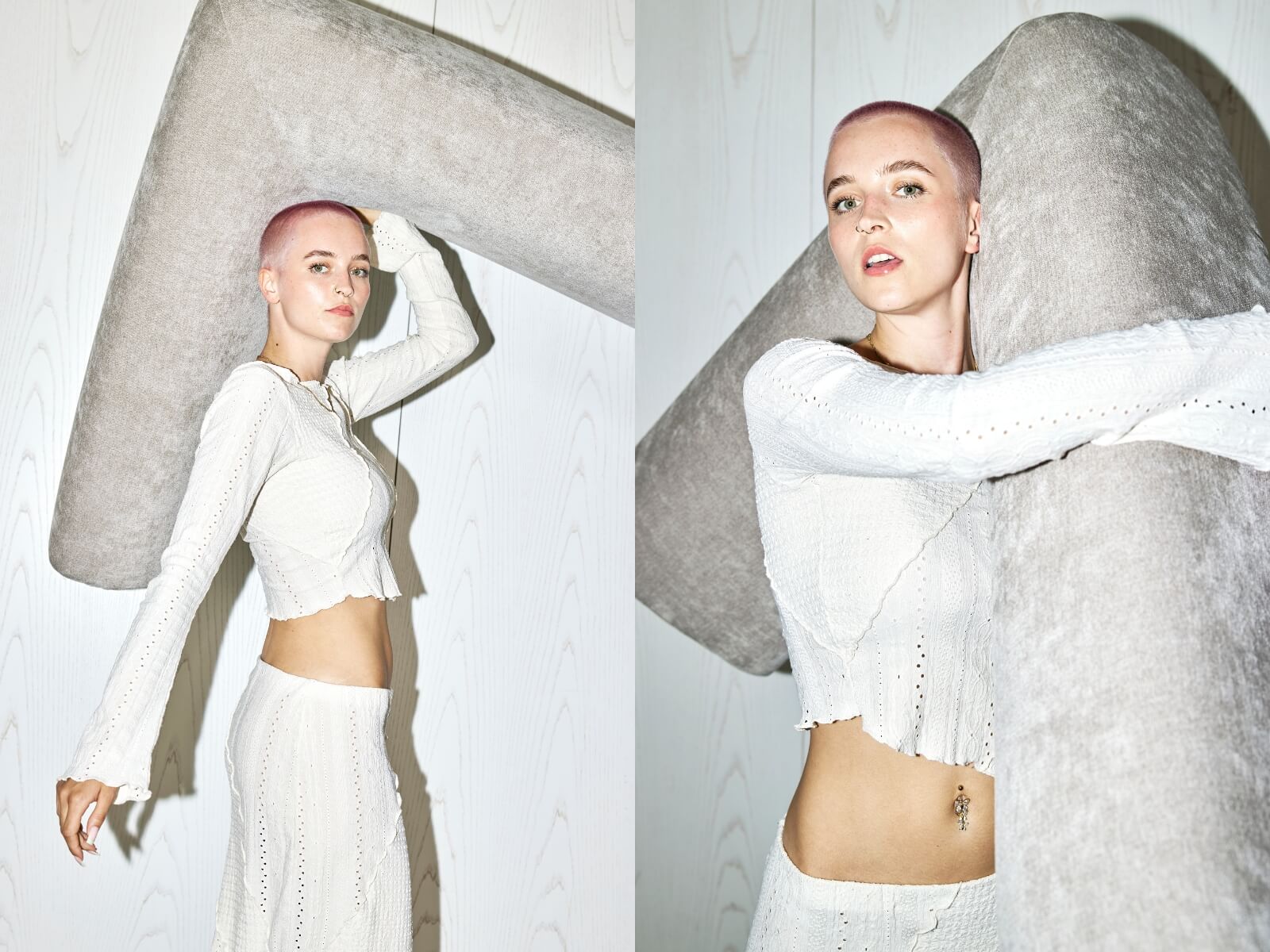 Eine Frau mit kurzen rosafarbenen Haaren, die ein weißes gehäkeltes Kleid trägt, posiert mit einem großen, weichen, hellgrauen L-förmigen Objekt vor einer weiß getäfelten Wand. Sie steht selbstbewusst da und blickt in die Kamera.