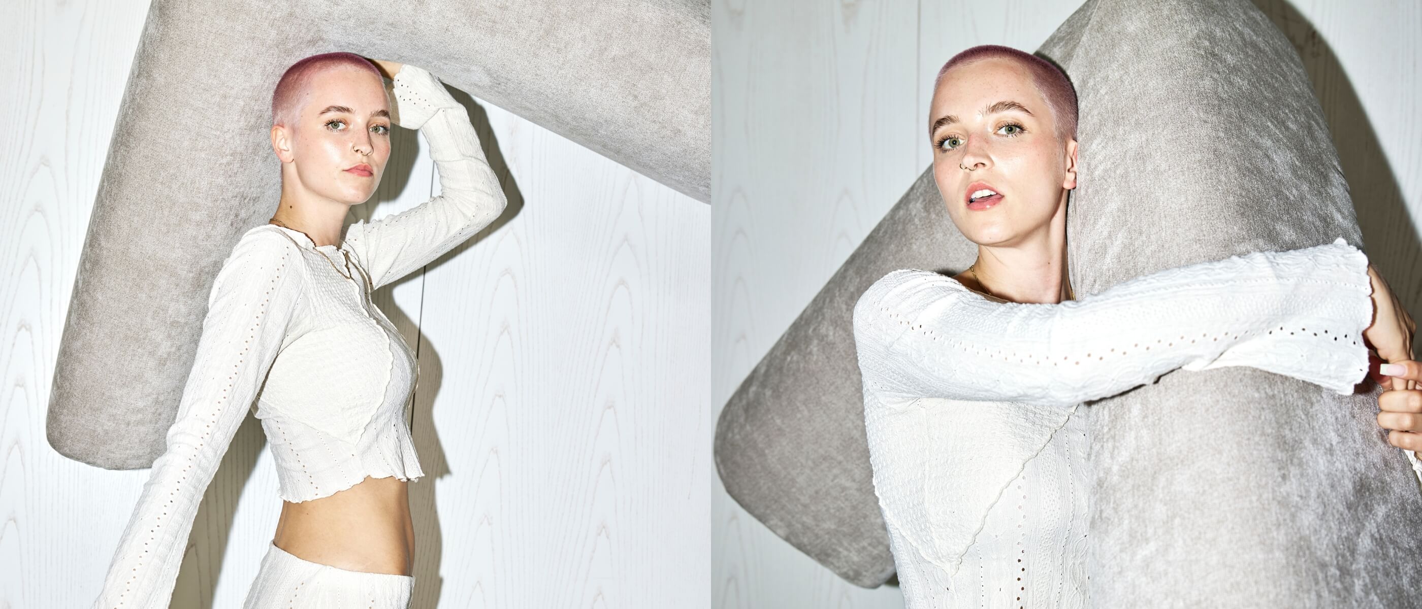 Eine Frau mit rasiertem Kopf und heller Kleidung posiert in einem Innenraum und lehnt sich an ein großes, weiches, hellgraues, L-förmiges Kissen an einer weißen Holzwand.