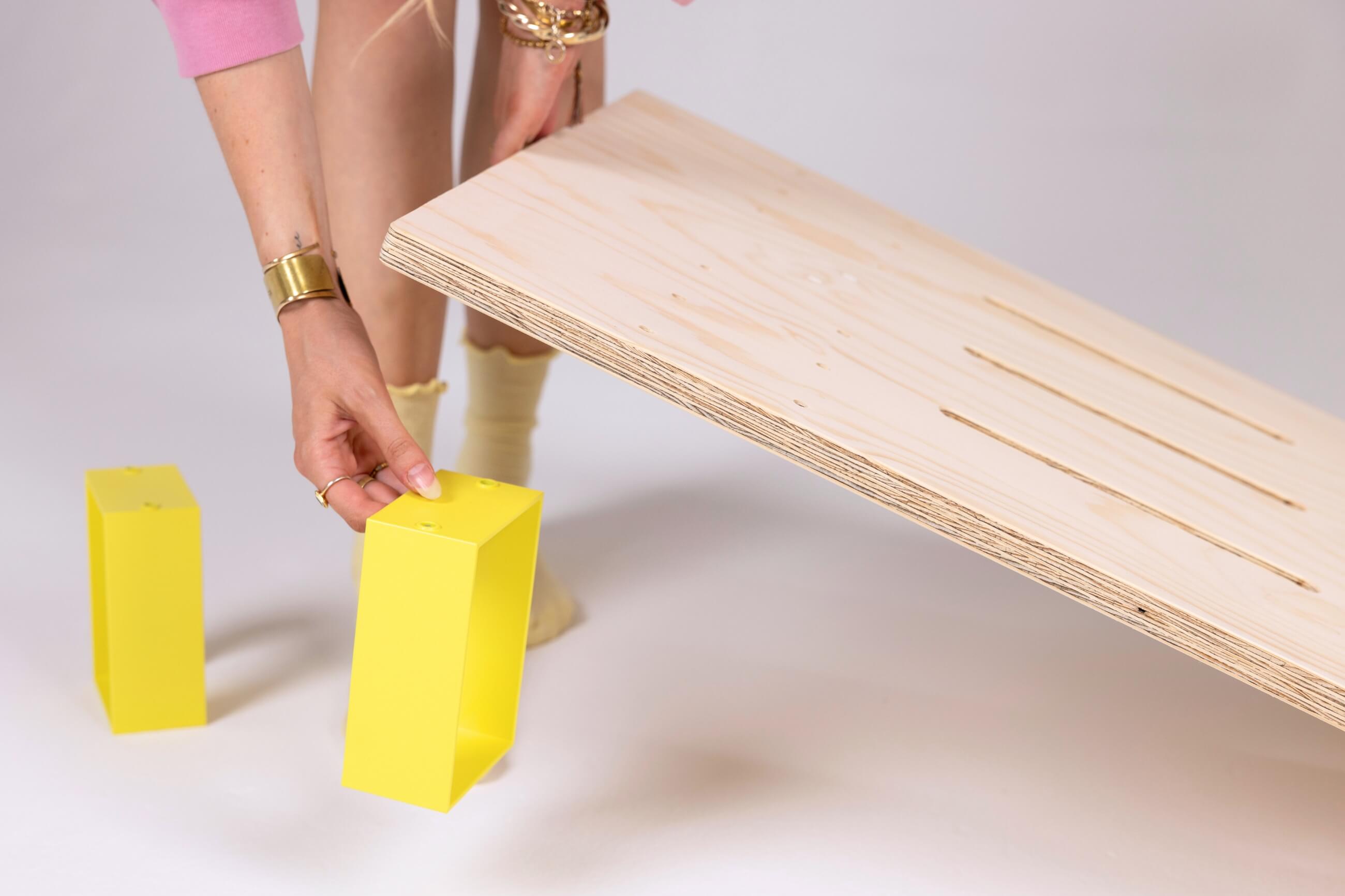 Eine Person in einem rosafarbenen Oberteil und mit Schmuck baut Möbel zusammen, hält ein Holzbrett in der Hand und legt einen gelben rechteckigen Block vor einem schlichten Hintergrund darunter.
