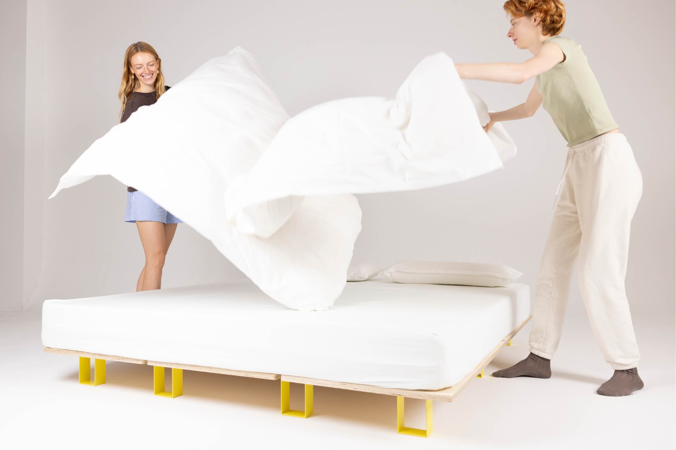Zwei Personen machen gemeinsam ein Bett, wobei sie eine weiße Bettdecke über ein ordentlich gemachtes Bett mit einem einfachen Holzrahmen und gelben Beinen in einem hellen, minimalistischen Raum ausbreiten.