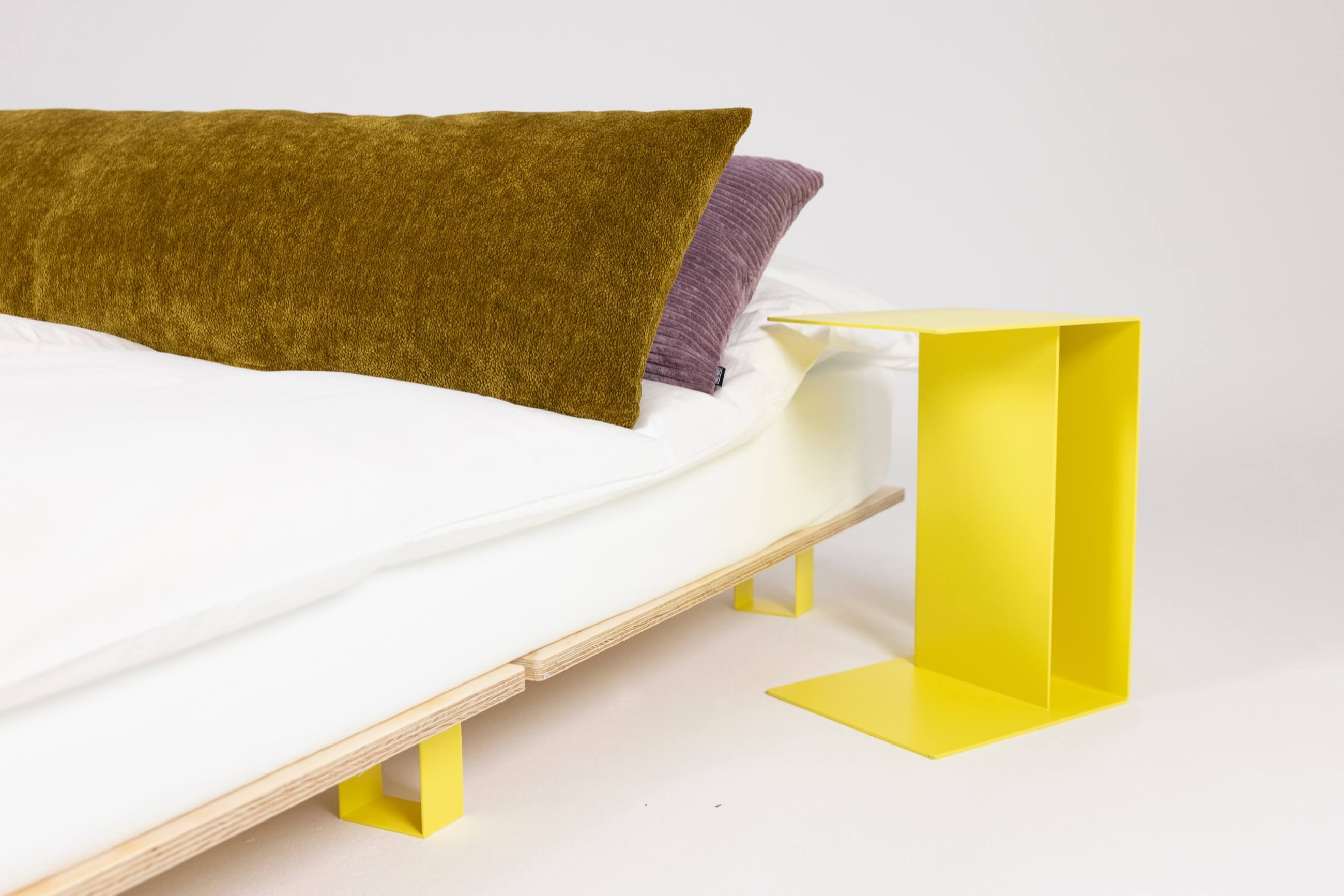 Ein modernes Bett mit weißer Bettwäsche, mit einem großen senfgelben Kissen und einem kleineren lila Kissen, neben einem minimalistischen hellgelben Nachttisch aus Metall. Die Szene hat einen klaren, zeitgenössischen Stil.