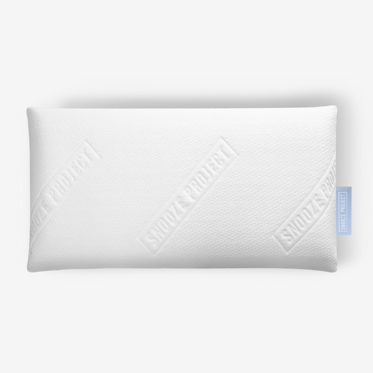 SPHVK_img002 Ein rechteckiges weißes Kissen mit strukturierter Oberfläche, auf dem der Schriftzug SNOOZE PROJECT diagonal über den Stoff geprägt ist, sowie ein kleines blaues Etikett am rechten Rand.