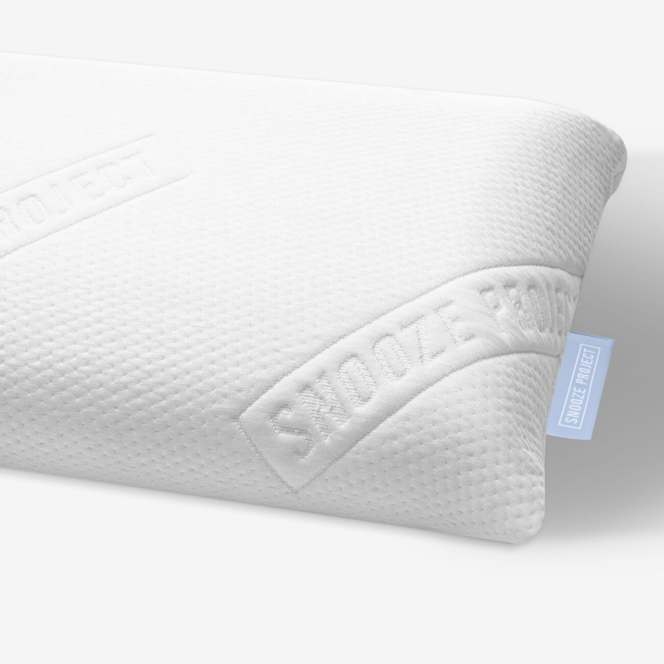 SPHVK_img003 Eine Nahaufnahme eines weißen, strukturierten Kissens mit der Prägung SNOOZE PROJECT auf dem Stoff. Ein kleines blaues Etikett mit weißem Text trägt ebenfalls die Aufschrift SNOOZE PROJECT. Der Hintergrund ist einfarbig weiß.
