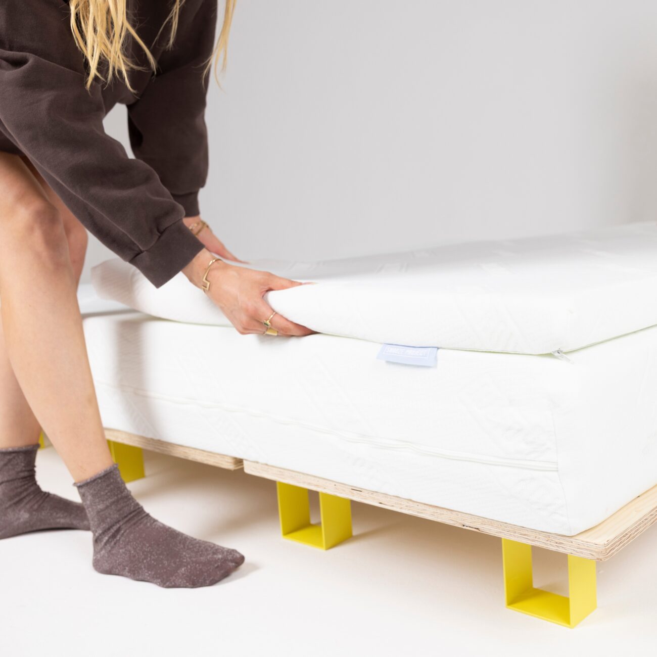 SPTFAM-Mood_img001 Eine Person, die graue Socken und einen dunklen Pullover trägt, hebt die Ecke einer weißen Matratzenauflage auf einem Bett mit einem einfachen Holzrahmen und gelben Beinen an.
